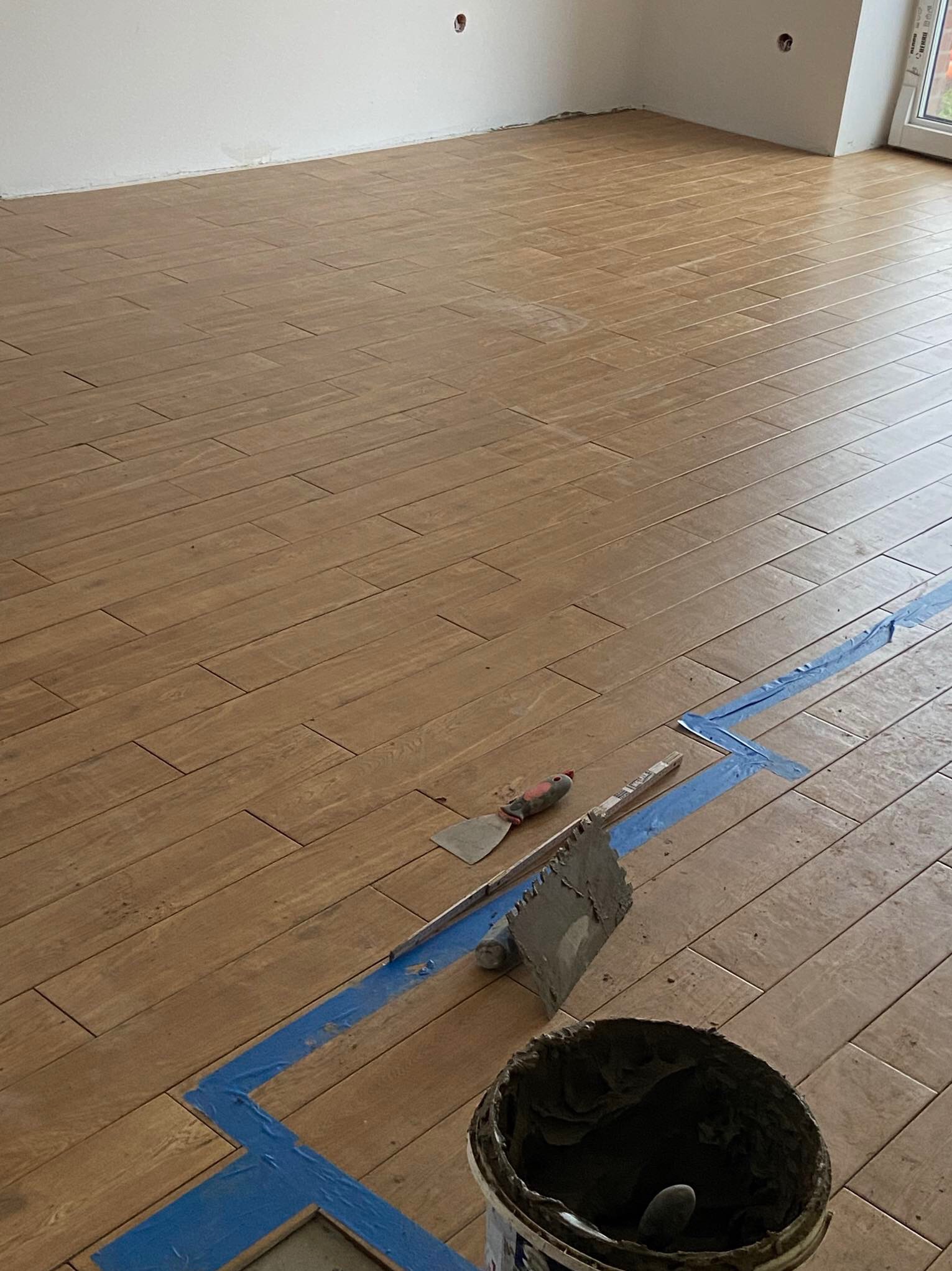 Układanie podłogi z płytek imitujących drewno, widoczne narzędzia: paca z zaprawą, szpachelka, poziomnica i wiadro z klejem, zabezpieczone taśmą malarską.