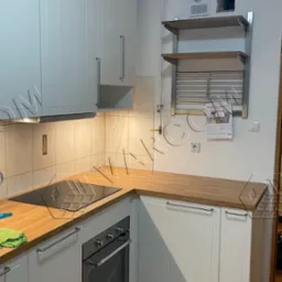 Biała kuchnia z drewnianym blatem, płytą indukcyjną i piekarnikiem, widoczne rury wentylacyjne i suszarka nad blatem