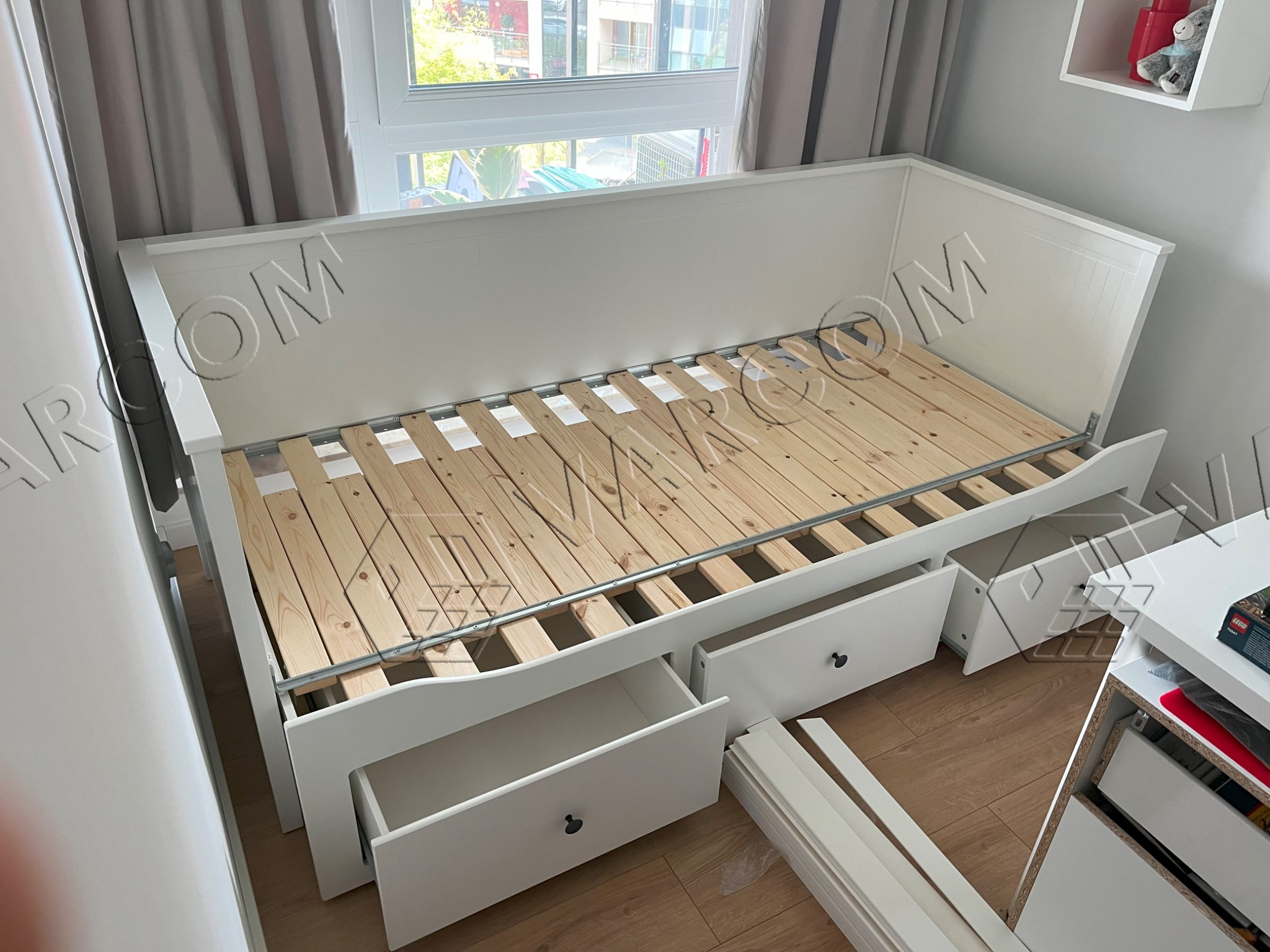 Biała rama łóżka z drewnianym stelażem i otwartymi szufladami, częściowo zmontowana, widoczne elementy montażowe na podłodze.