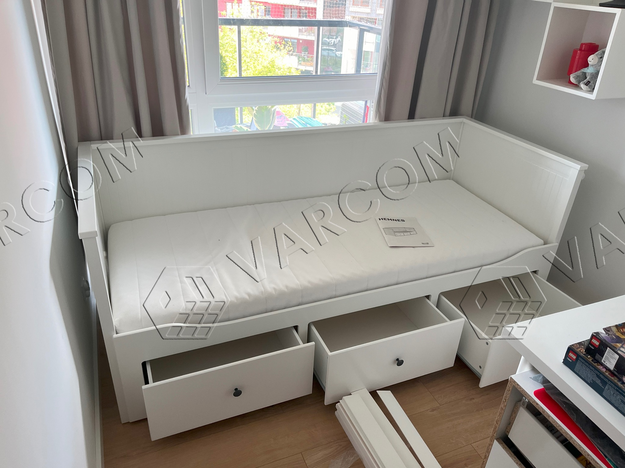 Białe łóżko dzienne z szufladami w pokoju dziecięcym, widok z przodu, częściowo otwarte szuflady, instrukcja obsługi na materacu, niedokończony montaż listew przypodłogowych.