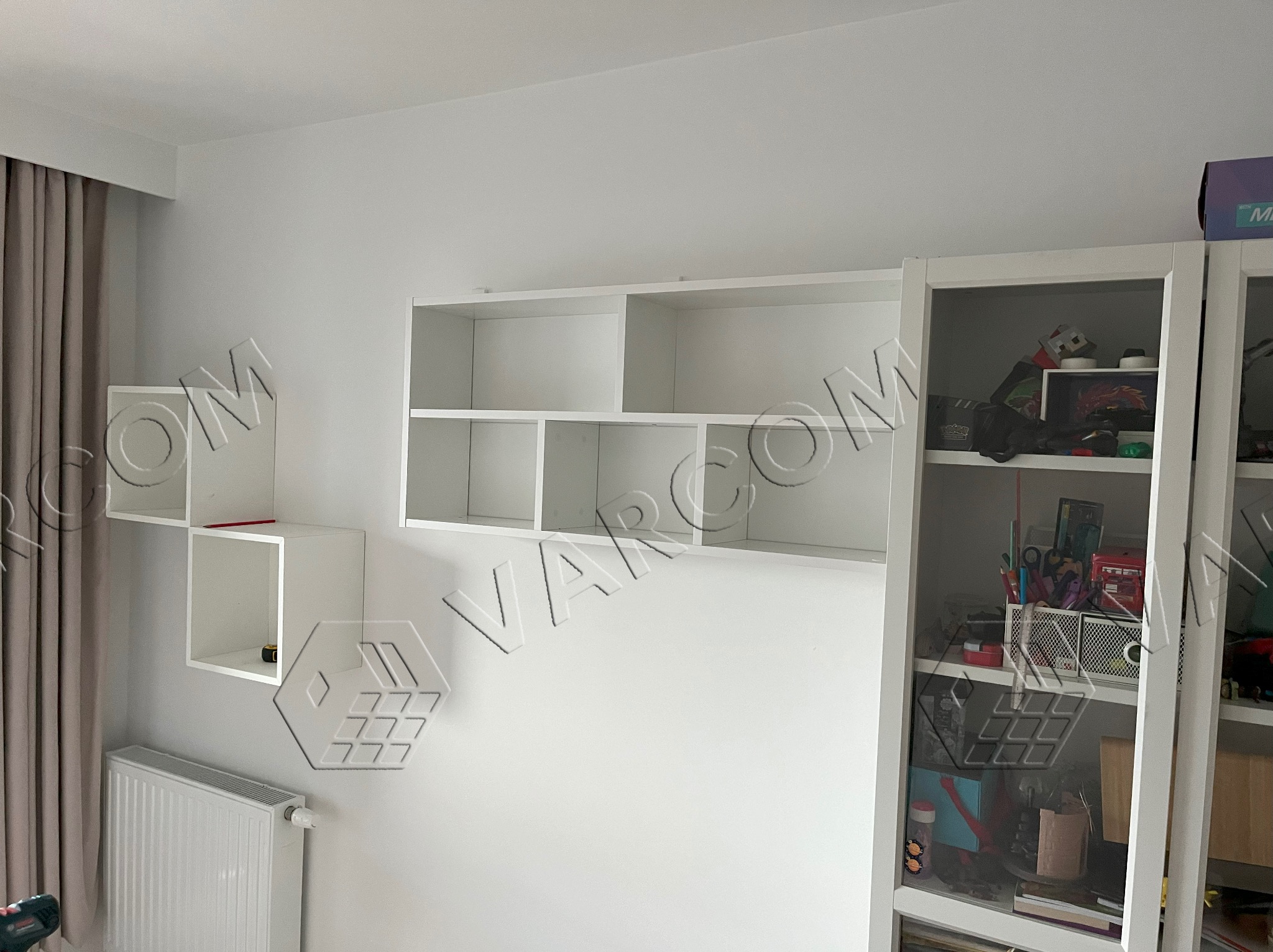 Białe, wiszące półki modułowe i regał z przeszklonymi drzwiami w pokoju, widoczny grzejnik.