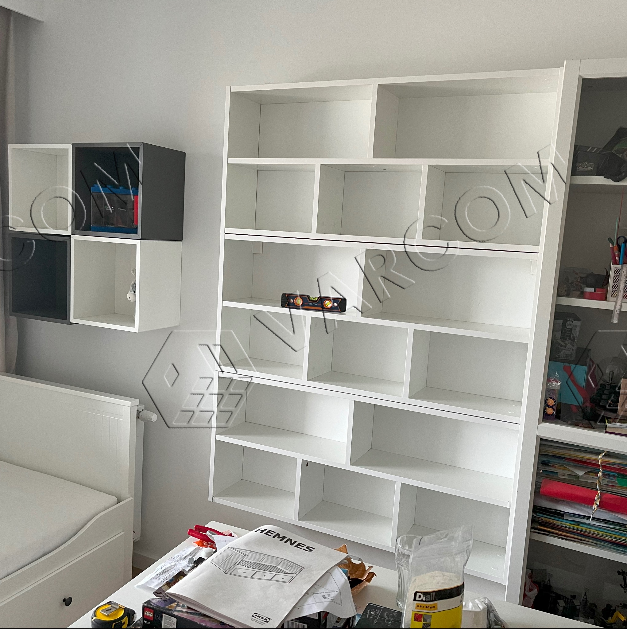 Biały regał z półkami, częściowo zamontowany, z poziomnicą na jednej z półek, instrukcją montażu Hemnes i narzędziami na blacie, obok łóżka.