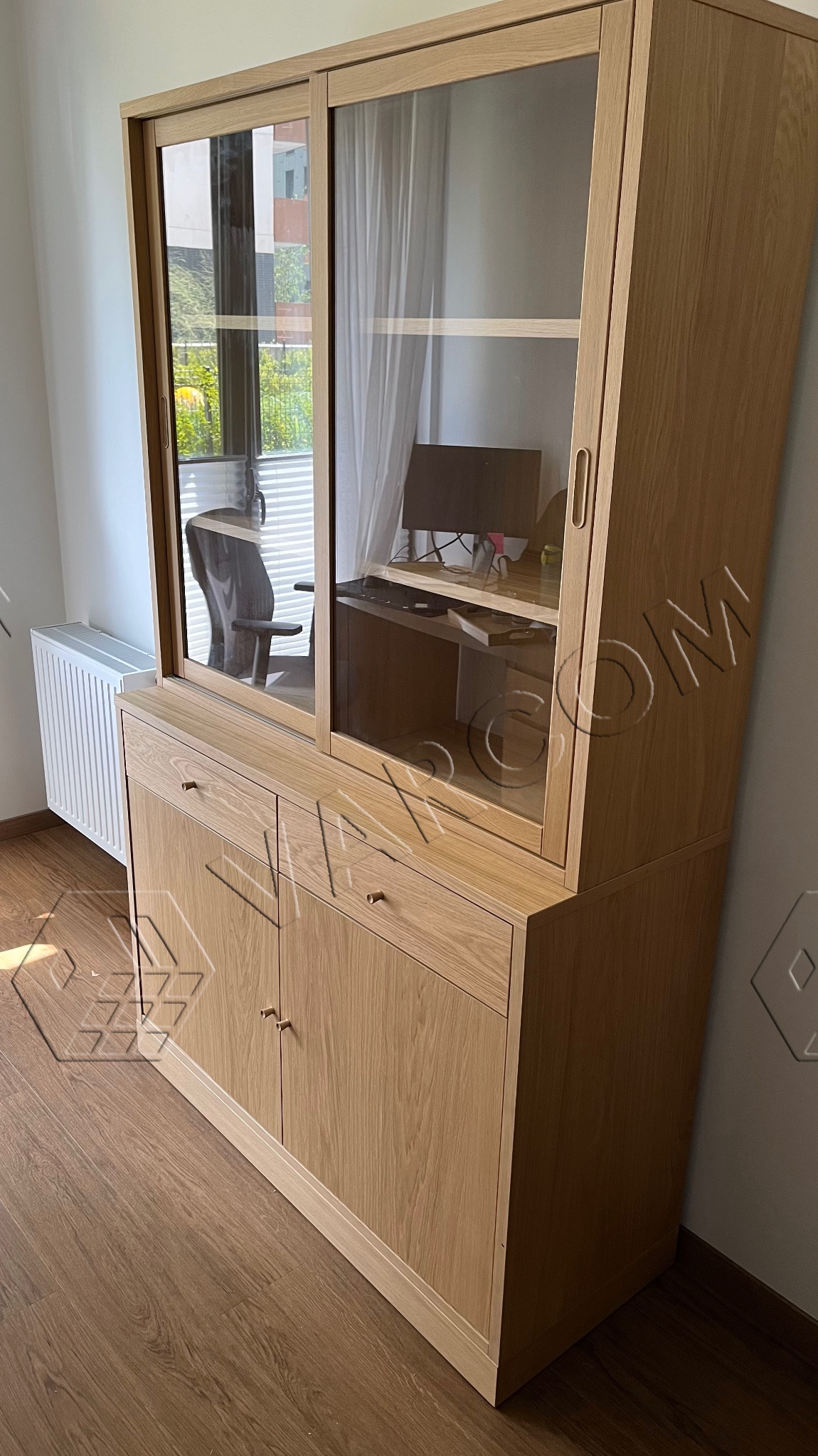 Drewniana witryna z przesuwnymi szklanymi drzwiami, wykonana na wymiar w Krakowie. Widoczne szuflady, szafki i półki, a w tle biurko z komputerem.