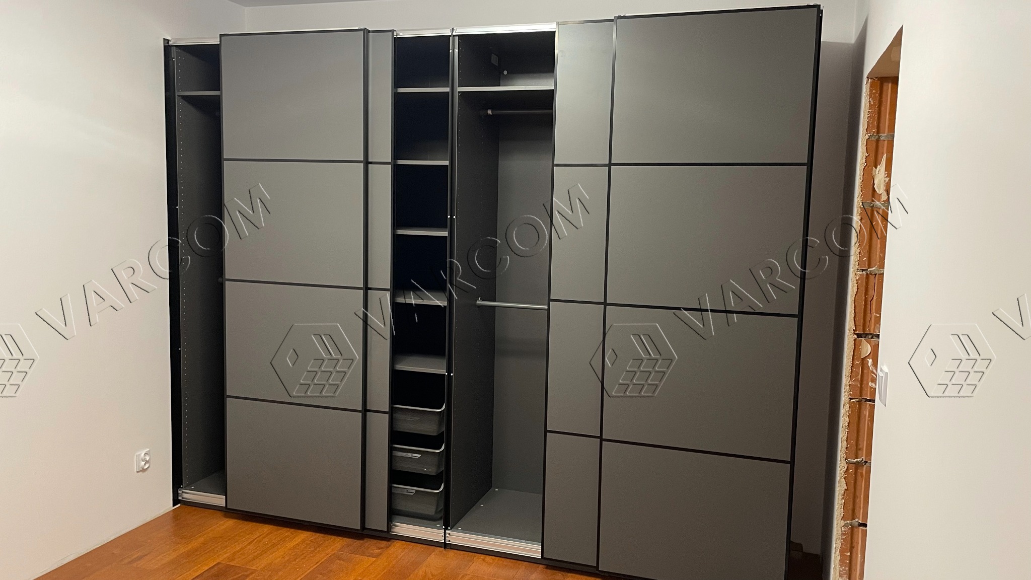 Szara szafa przesuwna z czarnymi obramowaniami, częściowo otwarta, ukazująca półki i drążek na ubrania, widoczna ściana z cegły w surowym stanie.