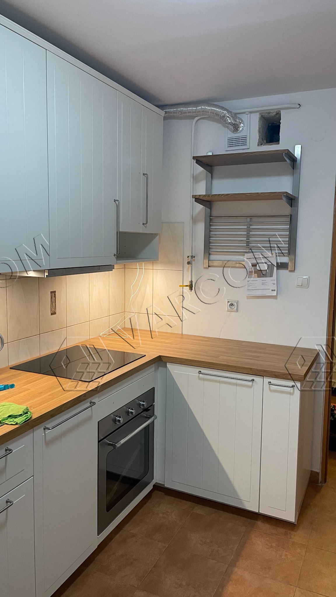 Biała kuchnia z drewnianym blatem, płytą indukcyjną i piekarnikiem, widoczne rury wentylacyjne i suszarka nad blatem