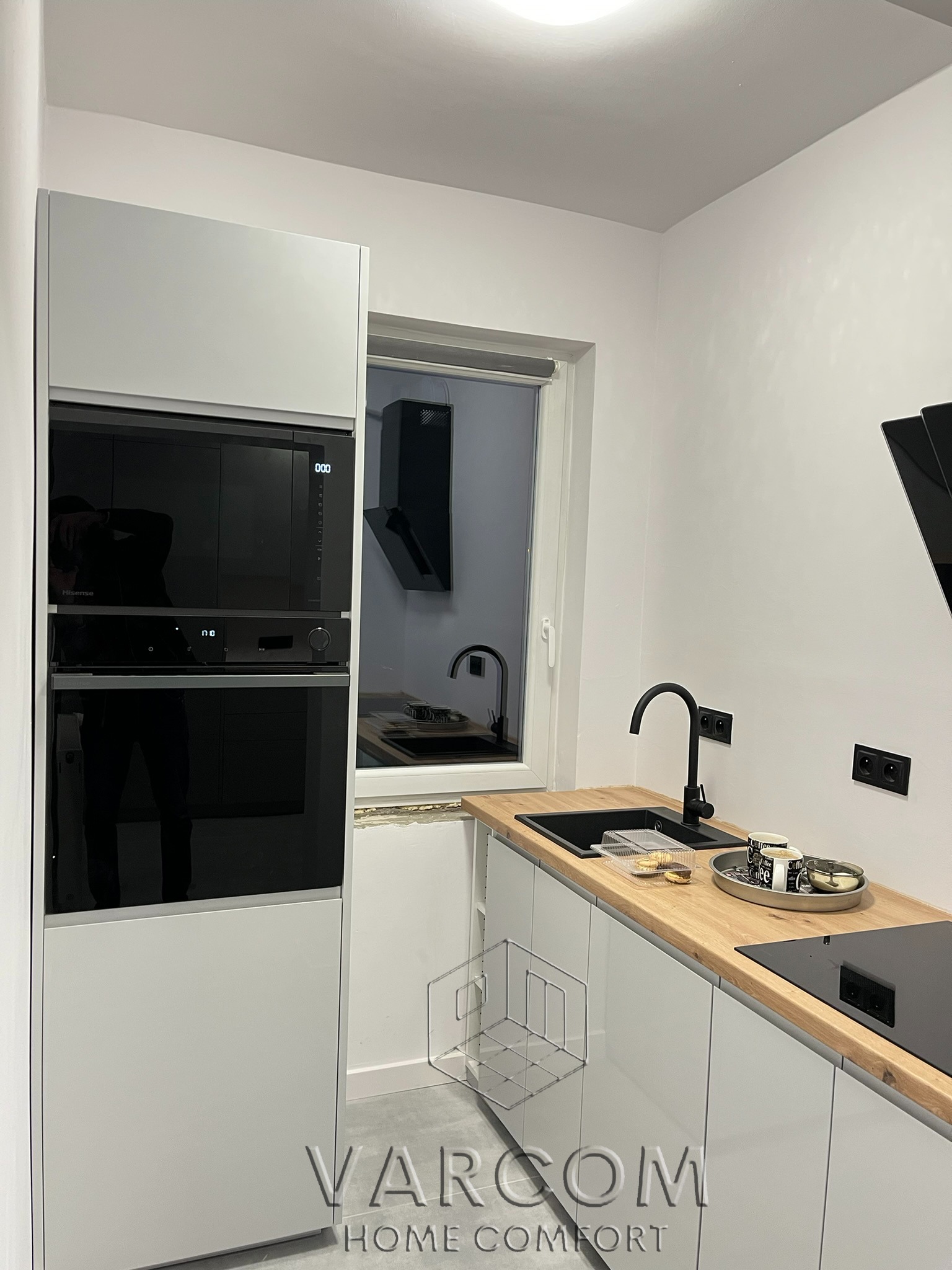 Nowoczesna kuchnia z białymi szafkami, drewnianym blatem i czarną armaturą. Widoczny piekarnik w zabudowie i okap. Na blacie naczynia i przekąski.