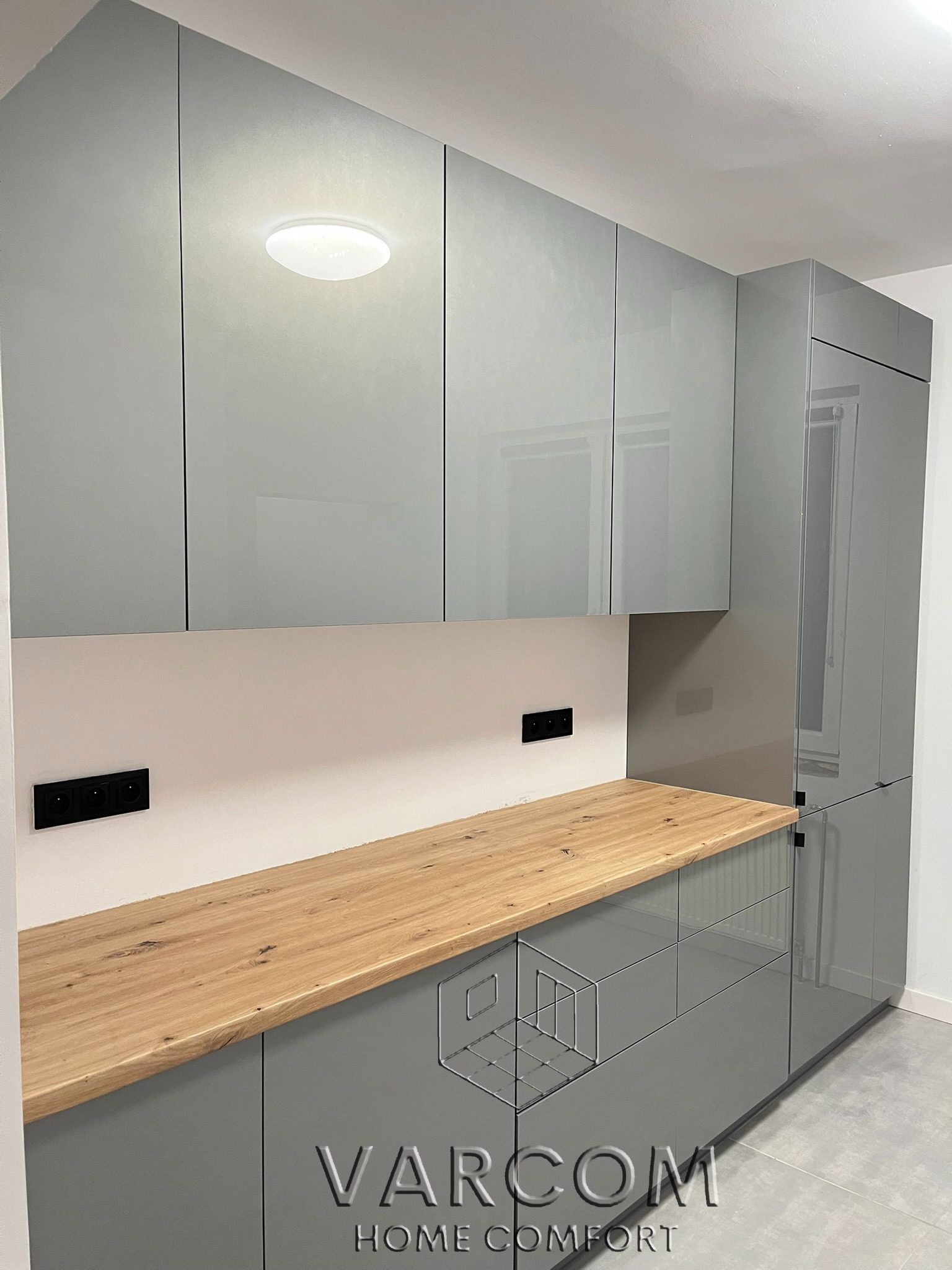 Szara, lakierowana zabudowa kuchenna z drewnianym blatem i czarnymi gniazdkami, oświetlona okrągłą lampą sufitową. Minimalistyczny design.