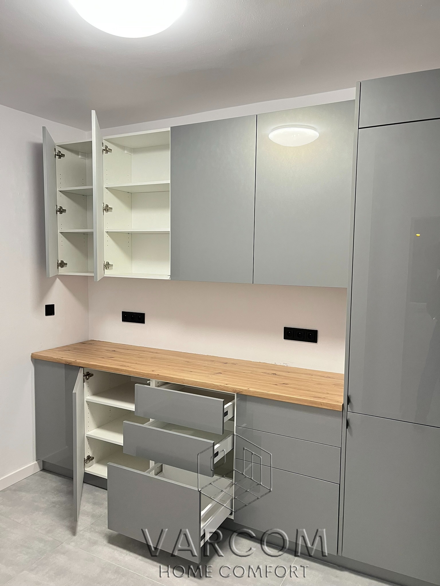 Szara zabudowa kuchenna z drewnianym blatem, otwartymi szafkami i wysuwanymi szufladami. Widoczne czarne gniazdka elektryczne na ścianie.