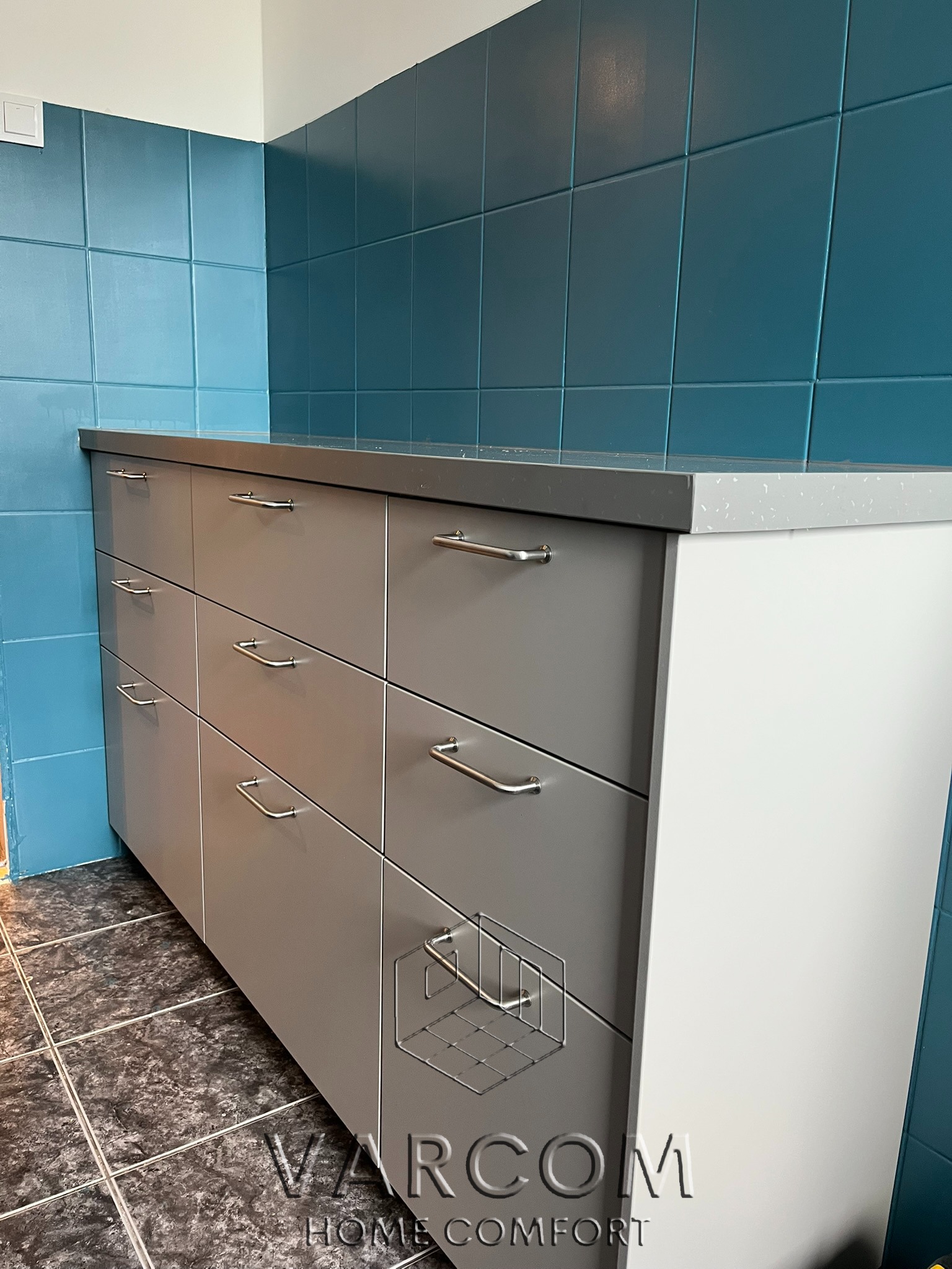 Szara szafka z sześcioma szufladami i srebrnymi uchwytami na tle turkusowych płytek. Blat szary z białymi plamkami. Widoczny fragment podłogi z ciemnych płytek.