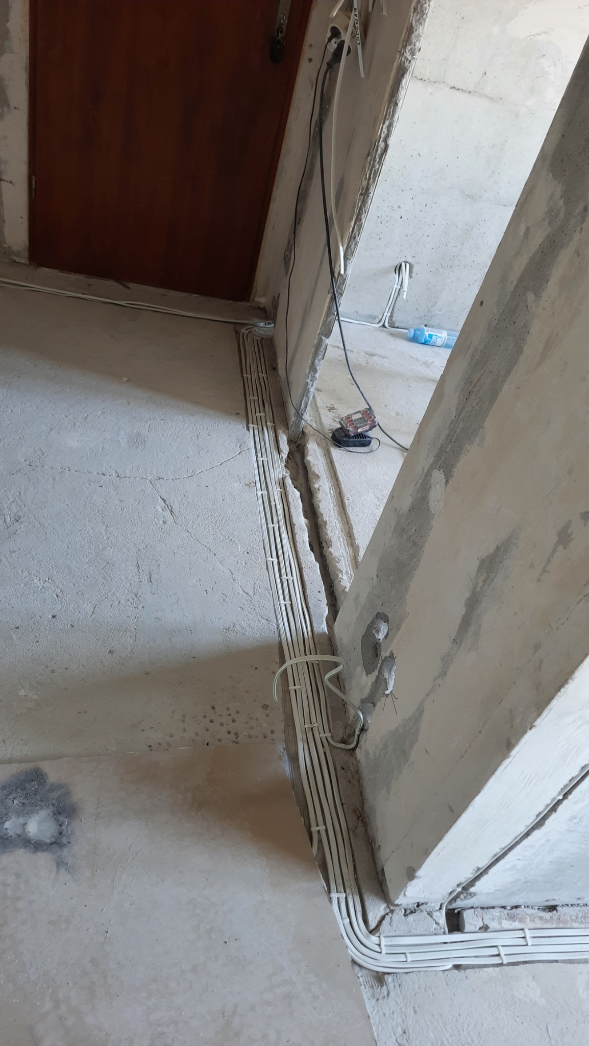 Białe przewody elektryczne ułożone w rzędzie wzdłuż surowej ściany podczas remontu, widoczny betonowy filar i fragment drzwi.