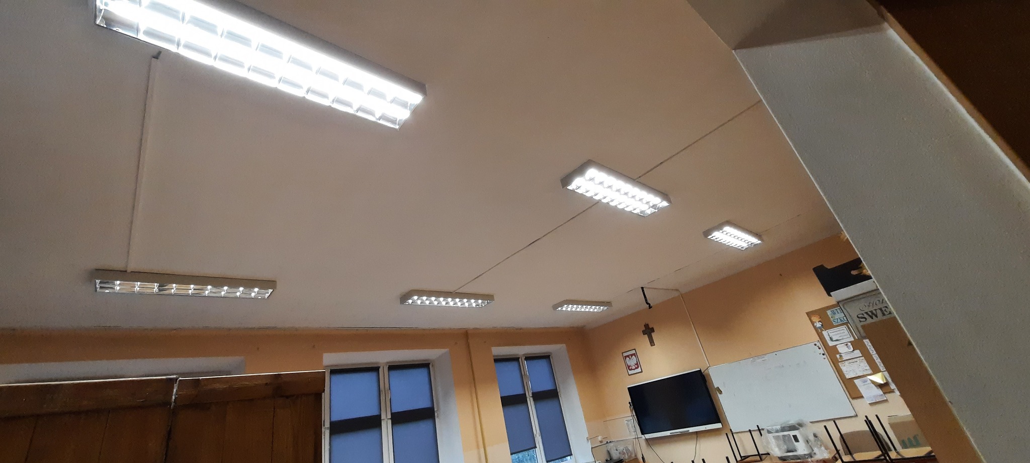 Równomiernie rozmieszczone, podłużne lampy sufitowe LED w jasnym pomieszczeniu, widoczne okna i tablica interaktywna na ścianie.