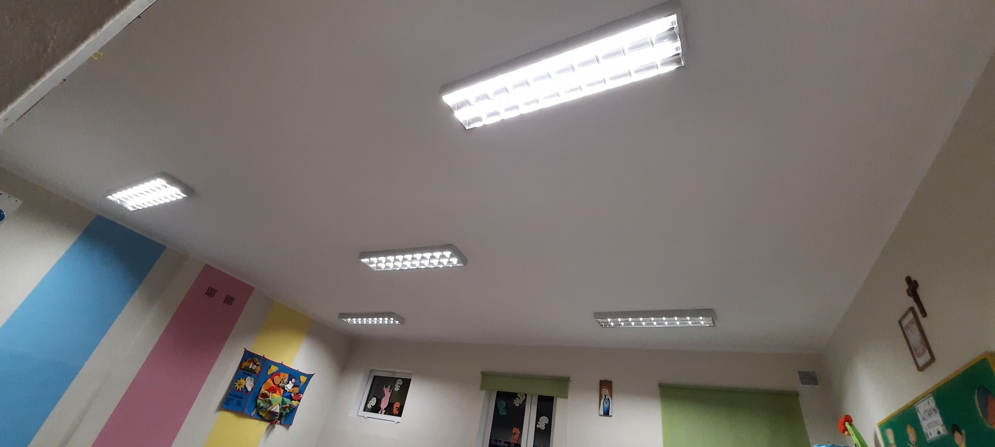 Sufit w przedszkolu z zamontowanymi lampami rastrowymi, widoczne kolorowe ściany i dekoracje okienne.