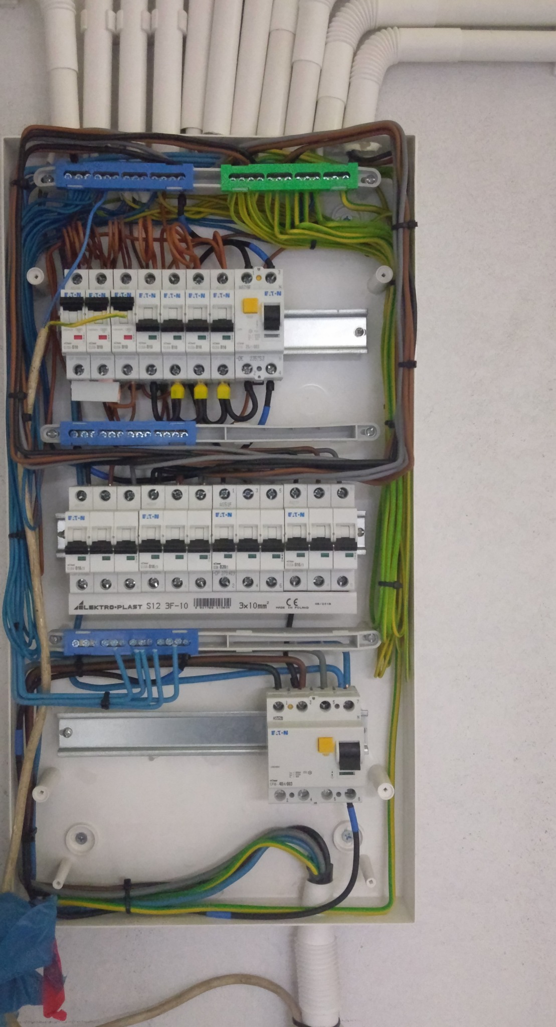 Otwarta skrzynka elektryczna z zainstalowanymi bezpiecznikami EATON i przewodami różnej grubości oraz kolorów, zamocowanymi na listwach zaciskowych wewnątrz białej obudowy.
