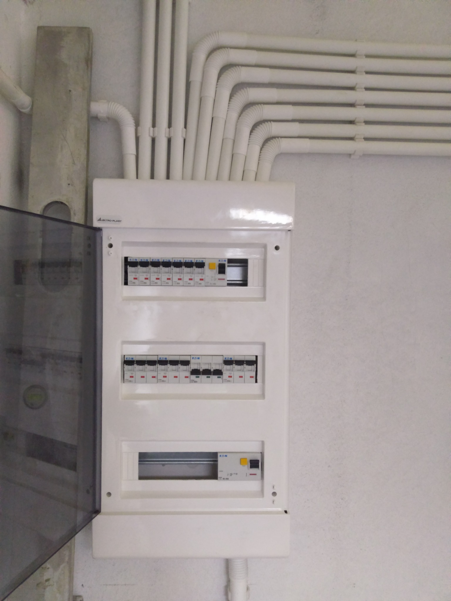 Rozdzielnica elektryczna firmy Elektro-Plast z otwartymi drzwiczkami, widoczne zabezpieczenia Eaton, białe rury PCV prowadzone po ścianie.
