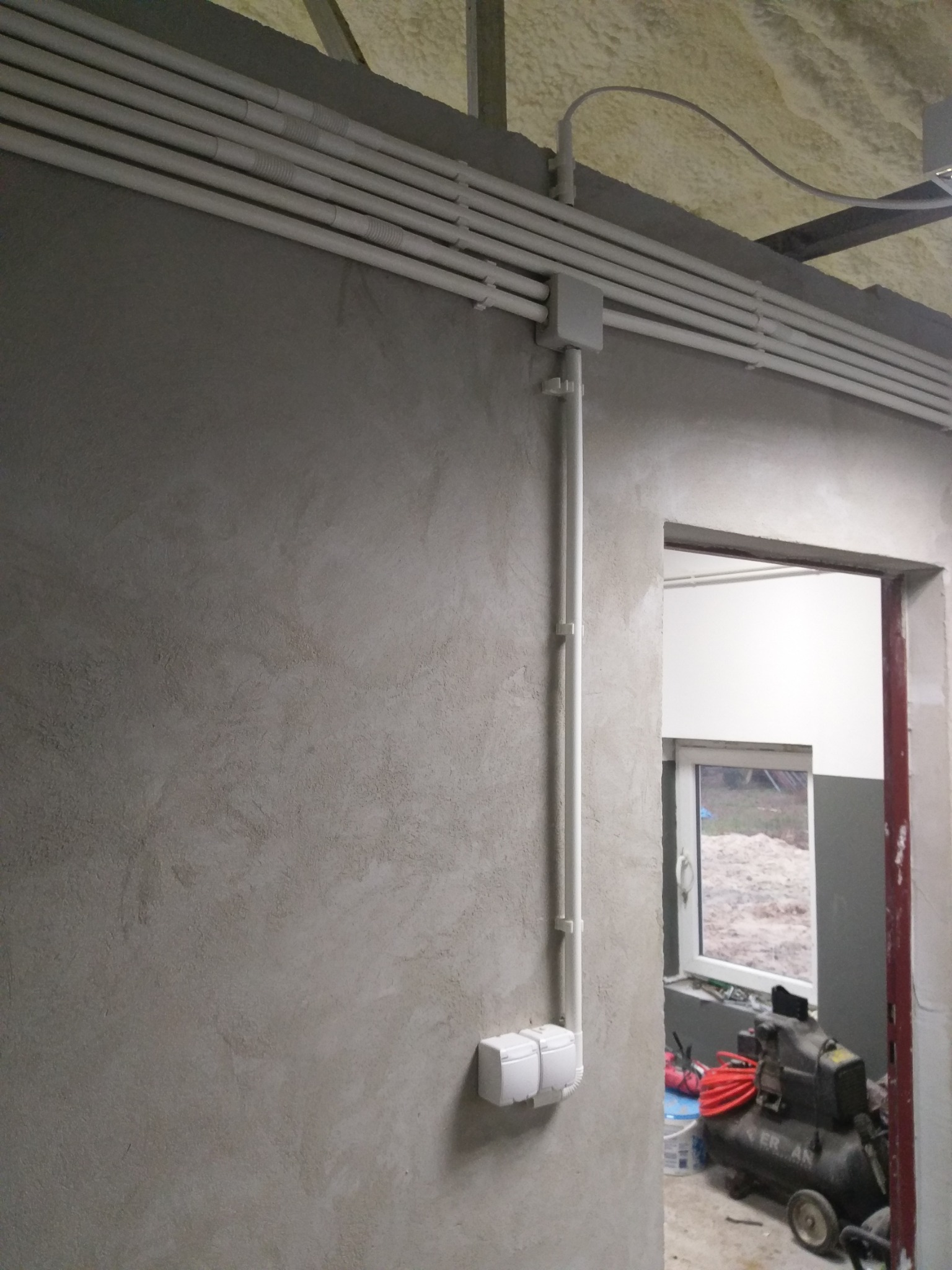 Szereg białych rurek PCV ułożonych równolegle pod sufitem, prowadzących wzdłuż ściany z gładzią, z puszką rozgałęźną i pionowym odcinkiem do podwójnego gniazdka elektrycznego; w tle widoczny...