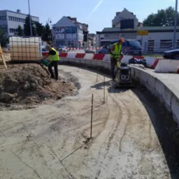 Dwóch robotników w kamizelkach odblaskowych podczas prac ziemnych: jeden ubija ziemię zagęszczarką, drugi polewa wodą z zielonego kanistra, w tle budynki miejskie i bariery drogowe.