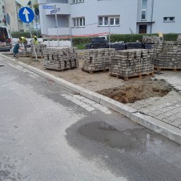M-Way Building - Układanie kostki brukowej na ulicy w dzień, widoczne palety z kostką, pracownicy w kamizelkach odblaskowych oraz zaparkowany samochód dostawczy z narzędziami.