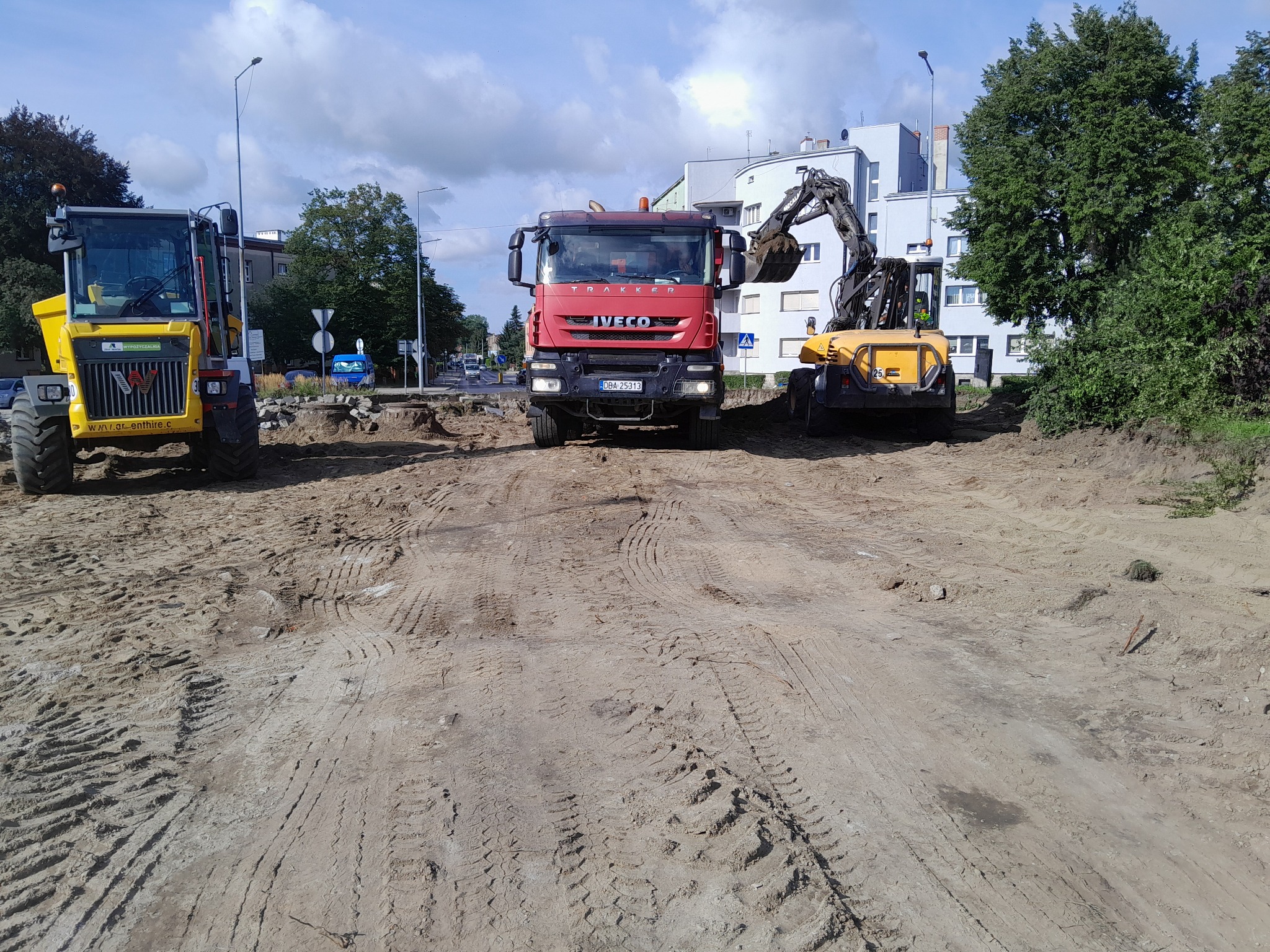 Ciężarówka IVECO i dwie maszyny budowlane na placu budowy z widocznymi śladami opon na piasku i ziemi, w tle budynki mieszkalne i drzewa.