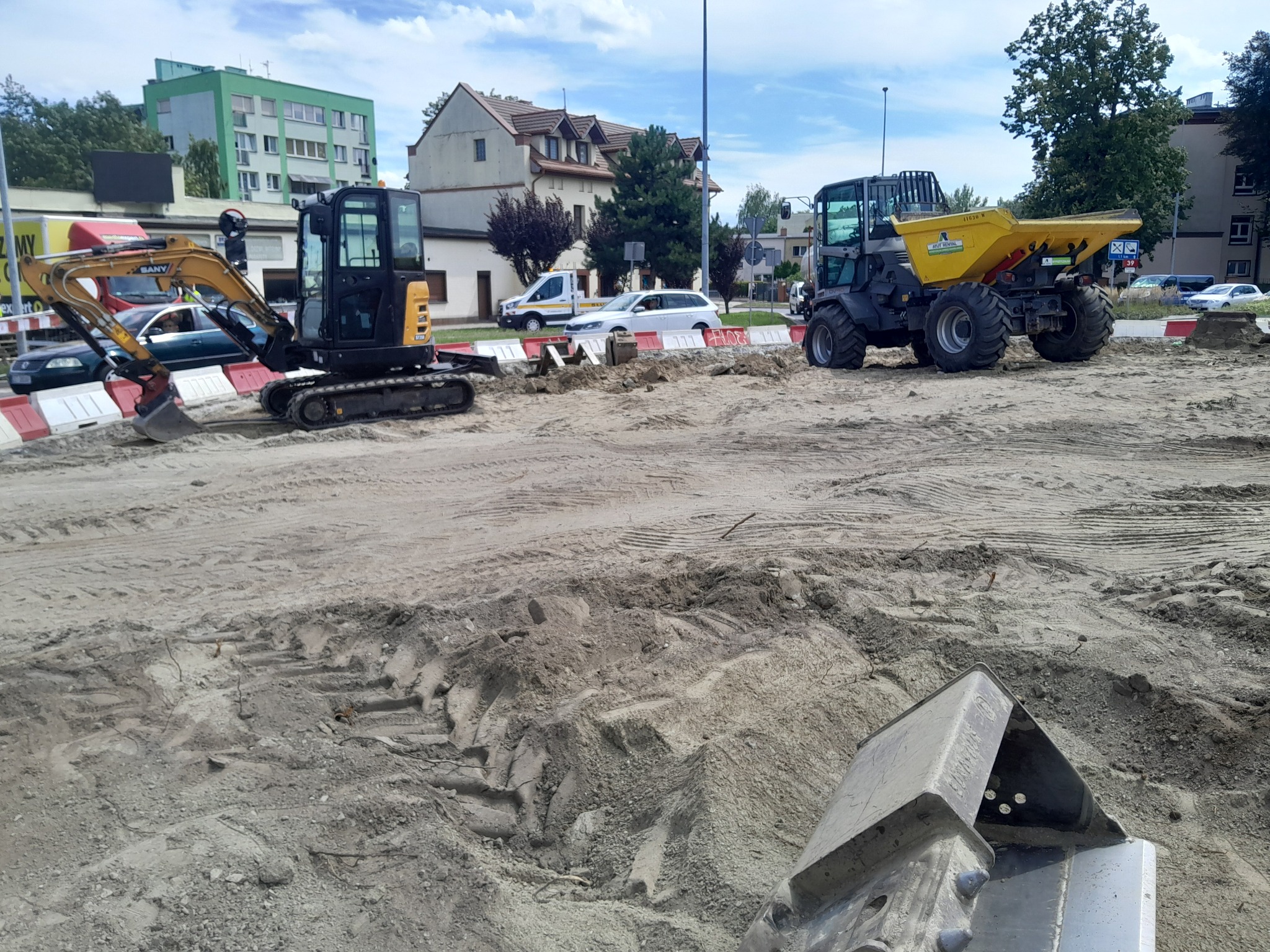 Dwa małe koparki i wywrotka na placu budowy z widocznymi śladami opon na ziemi, w tle budynki mieszkalne i samochody.