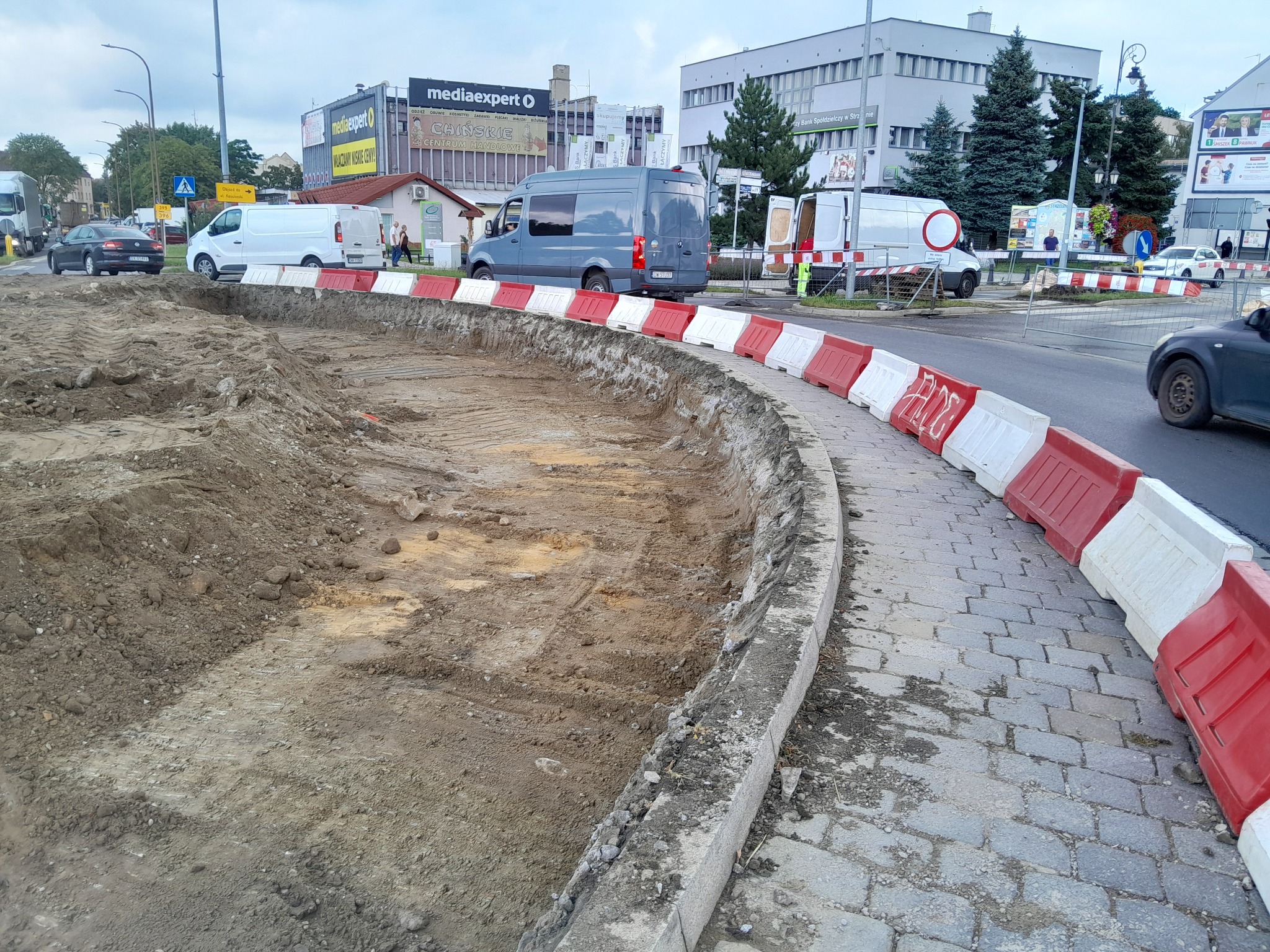 Przygotowany teren pod budowę, odgrodzony barierami z tworzywa sztucznego, widoczne ślady po oponach maszyn budowlanych, w tle budynki miejskie i zaparkowane samochody.