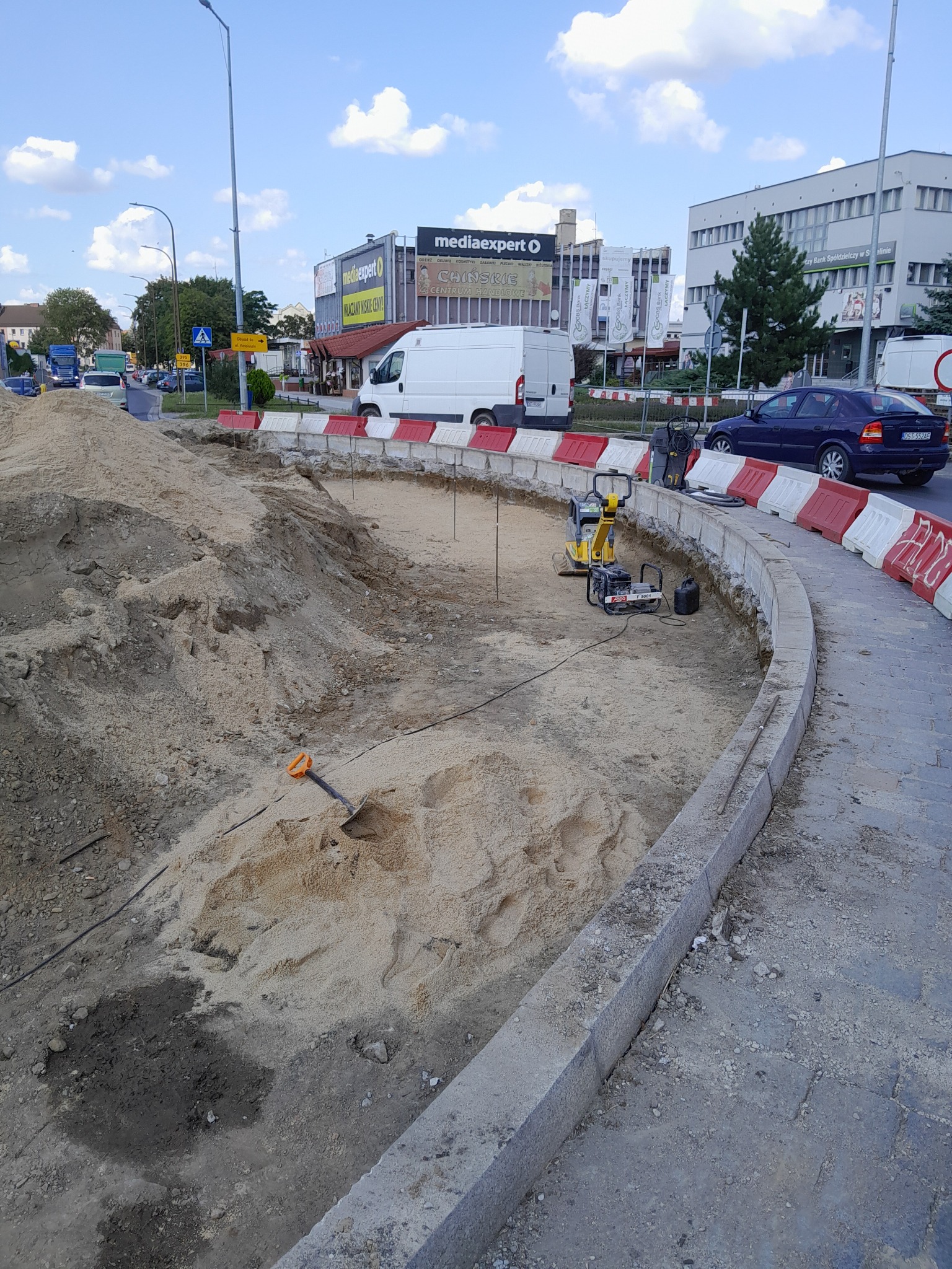 Przygotowanie terenu pod budowę ronda: wykop wypełniony piaskiem, ubijarka spalinowa, łopata z pomarańczową rączką, krawężniki zaporowe, budynki w tle.