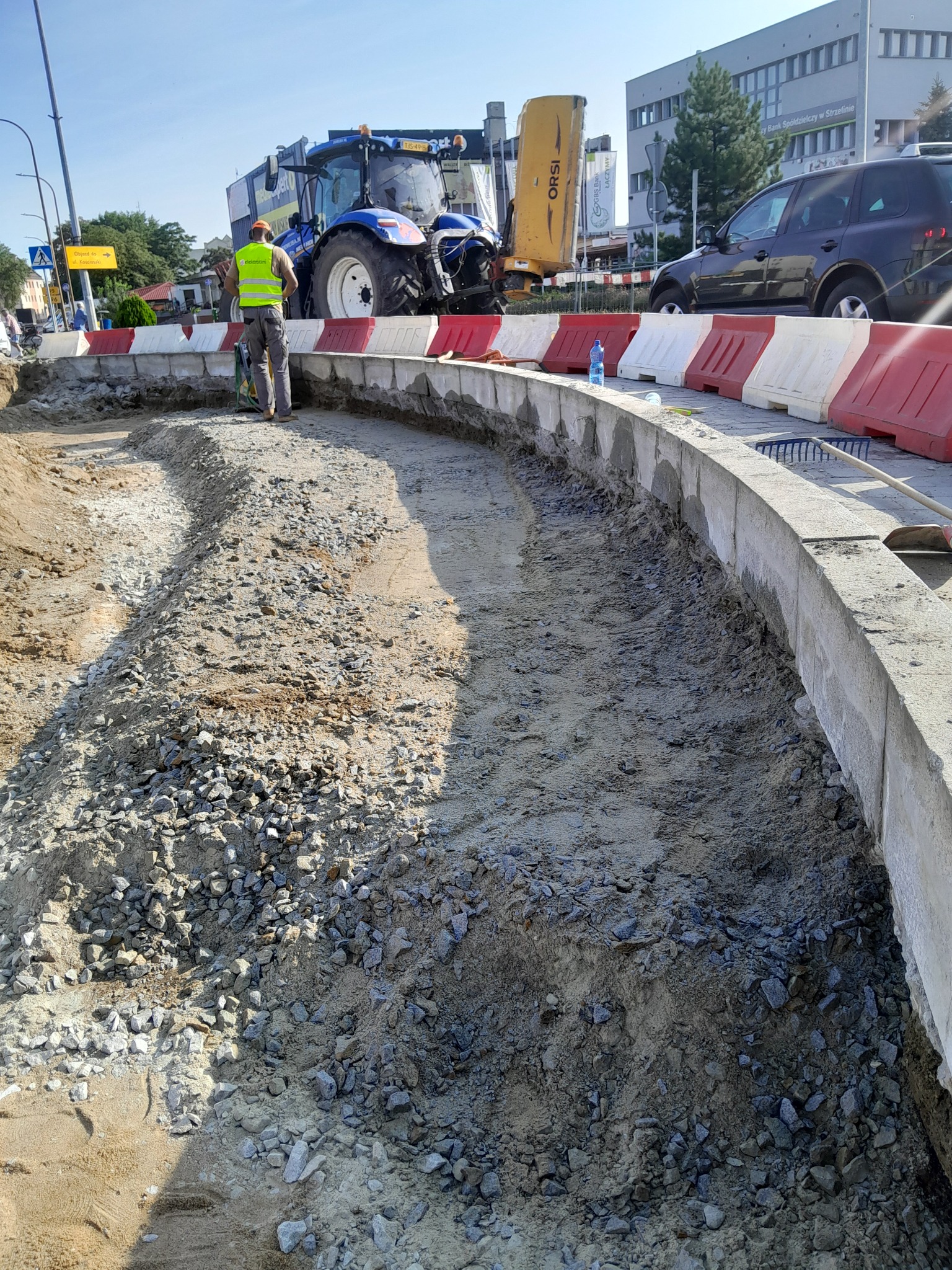 Przygotowanie podłoża pod chodnik z użyciem traktora i ubijarki, widoczny pracownik w kamizelce odblaskowej, krawężniki, piasek i kamień, w tle budynki miejskie.