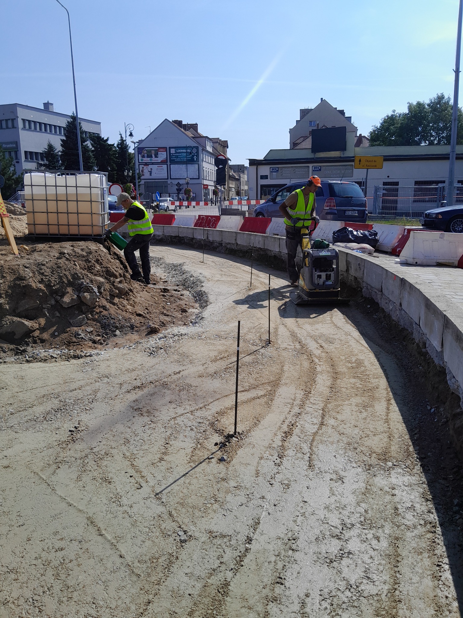 Dwóch robotników w kamizelkach odblaskowych podczas prac ziemnych: jeden ubija ziemię zagęszczarką, drugi polewa wodą z zielonego kanistra, w tle budynki miejskie i bariery drogowe.