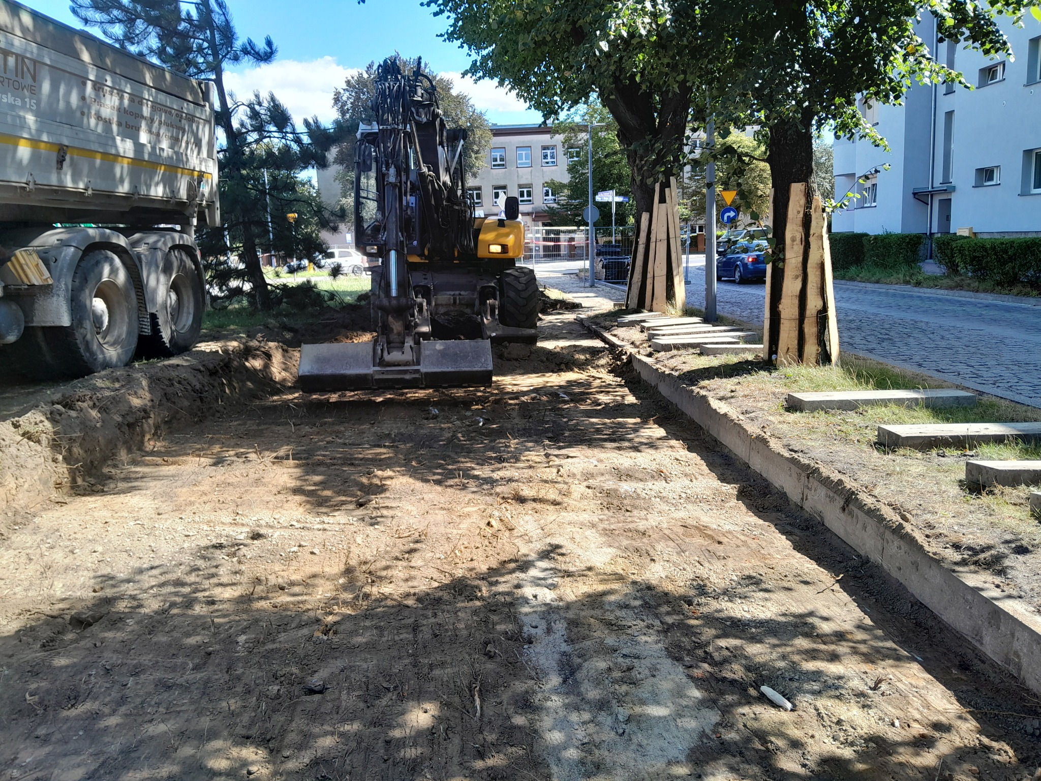 Żółta koparka kołowa w trakcie wykonywania wykopu pod drogę, widoczny fragment ciężarówki z piaskiem i zabezpieczone pnie drzew na tle miejskiej ulicy.