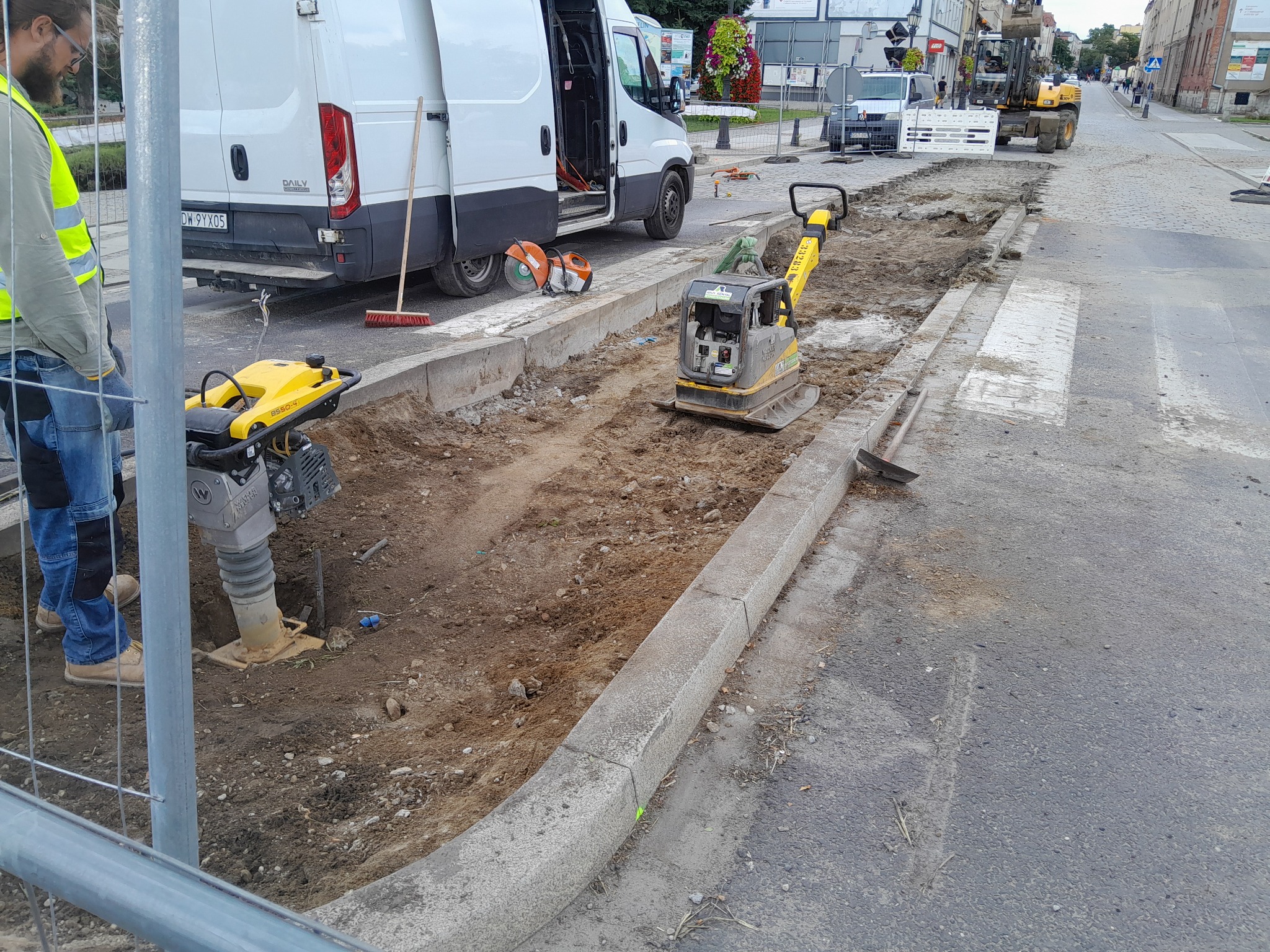 Przygotowanie podłoża pod nową nawierzchnię drogi: wykop koryta, ubijarki w akcji, biały bus z otwartymi drzwiami oraz pracownik w odblaskowej kamizelce w tle miejskiej ulicy.