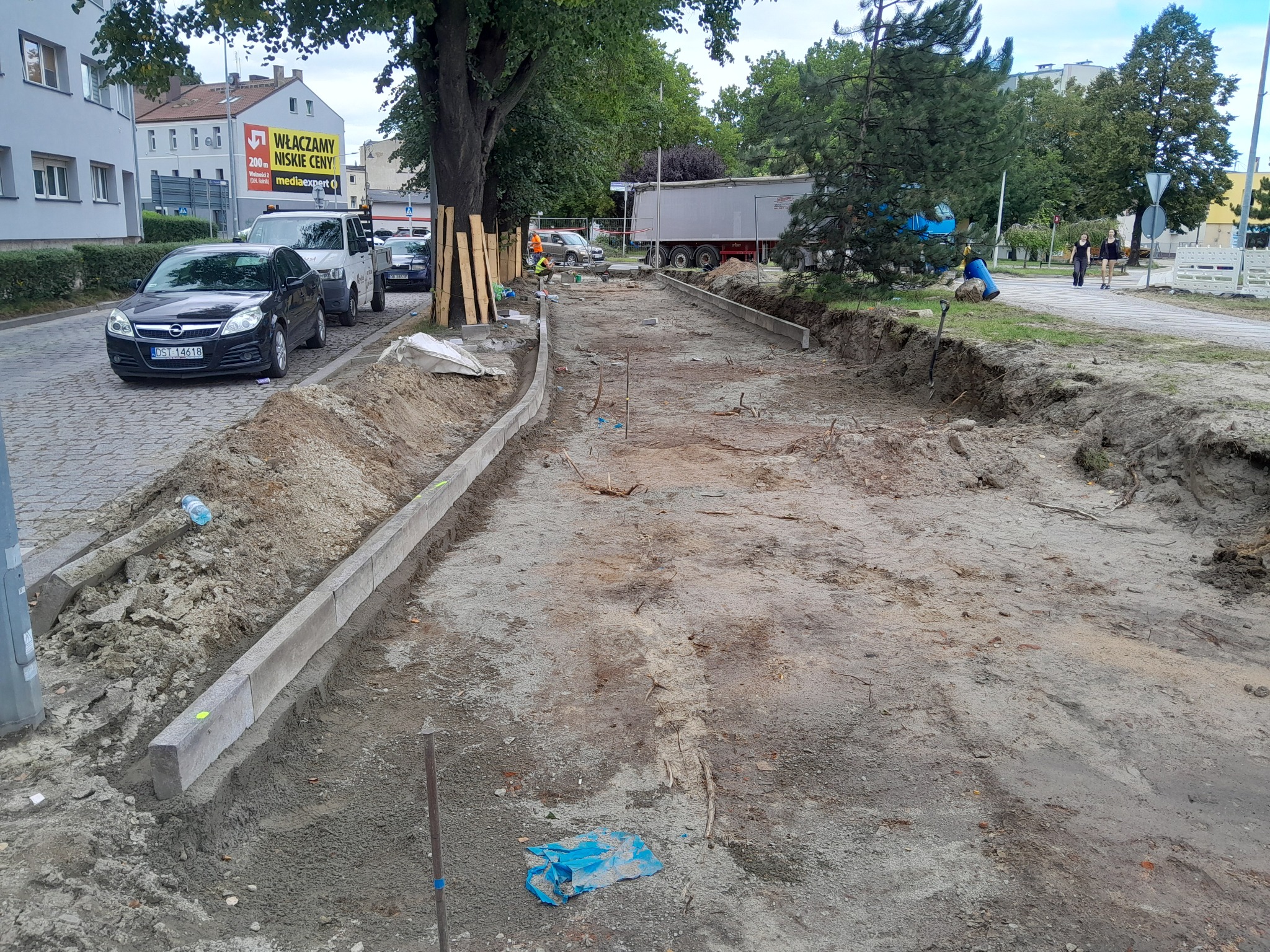 Przygotowanie terenu pod chodnik z ułożonymi krawężnikami, widoczne wykopane rowy, piasek i częściowo ułożone betonowe obrzeża, na drugim planie pracownicy w kamizelkach odblaskowych i zaparkowane...