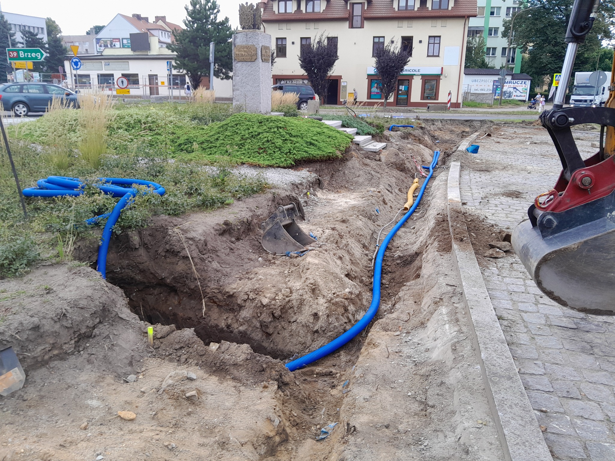 Głęboki wykop w ziemi z ułożonymi niebieskimi rurami, fragment koparki z łyżką widoczny po prawej stronie, w tle zieleń miejska i budynki w Brzegu.