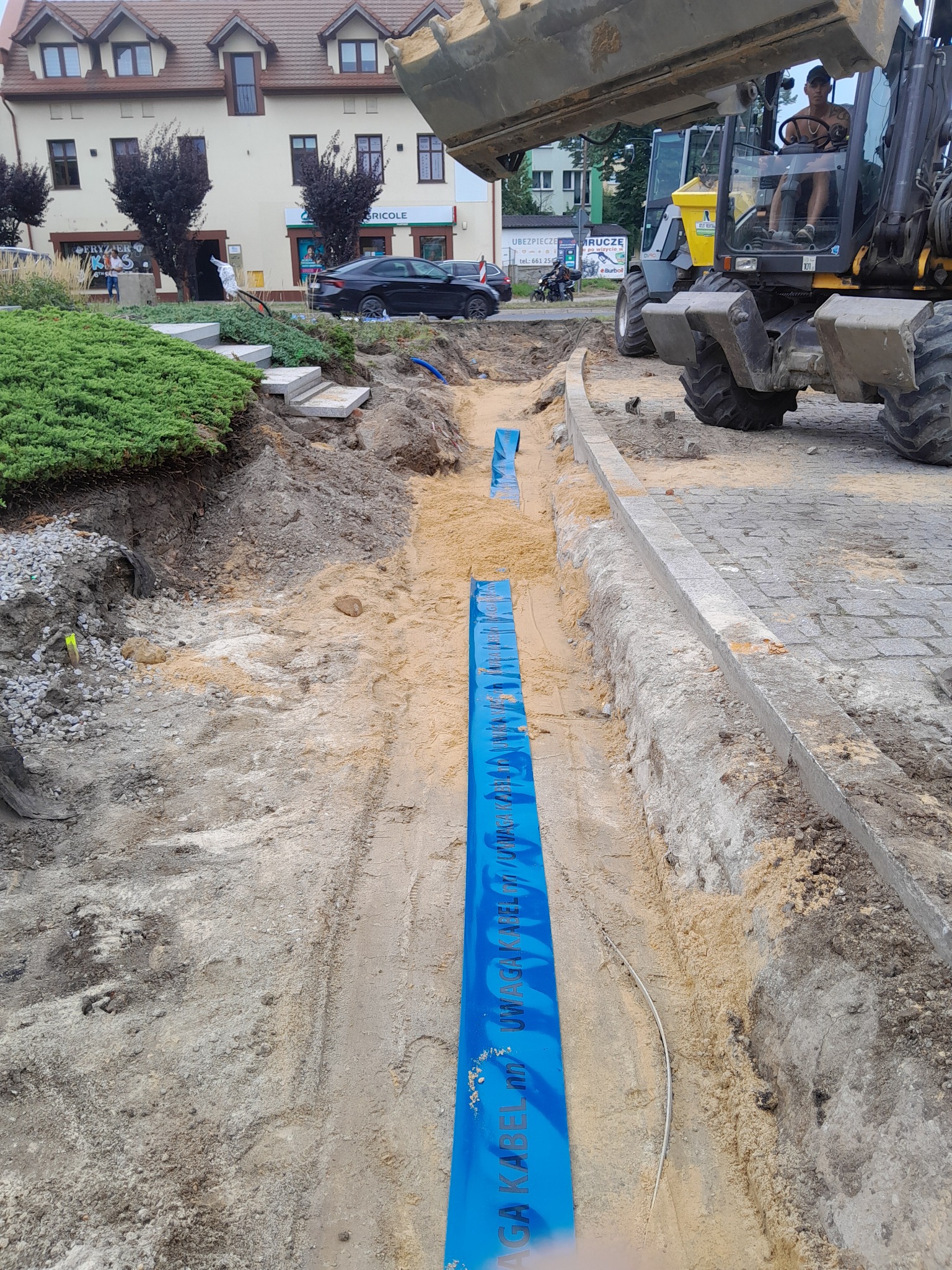 Wykopany rów z ułożonym niebieskim kablem w piasku, widoczna koparka i budynek w tle.