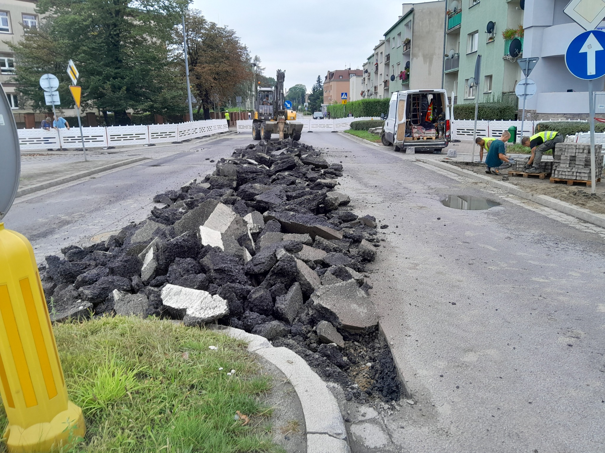 Fragment rozkopanej ulicy z gruzem asfaltowym, w tle ekipa robotników układająca kostkę brukową, widoczna maszyna budowlana i samochód dostawczy.