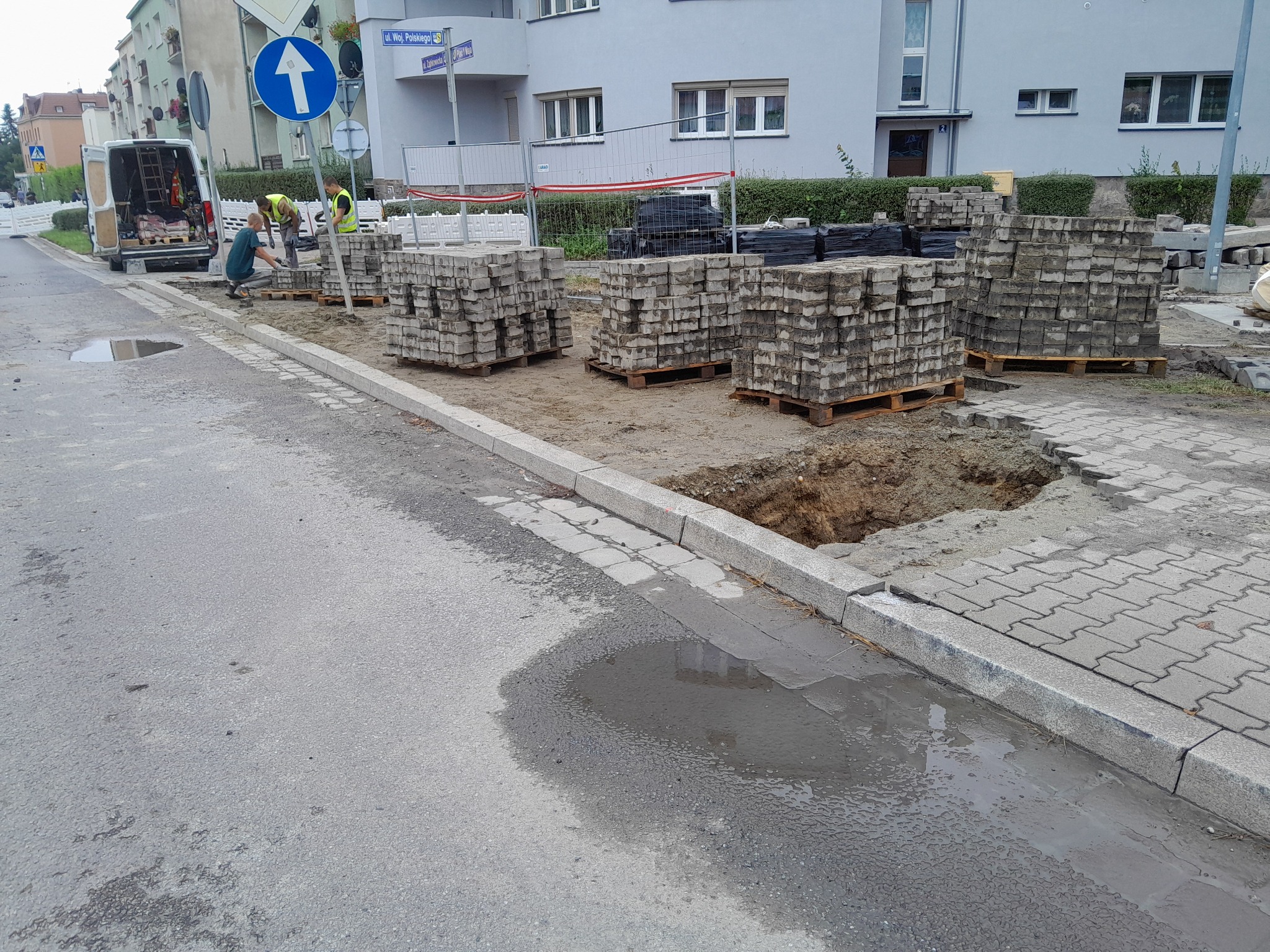 Układanie kostki brukowej na ulicy w dzień, widoczne palety z kostką, pracownicy w kamizelkach odblaskowych oraz zaparkowany samochód dostawczy z narzędziami.