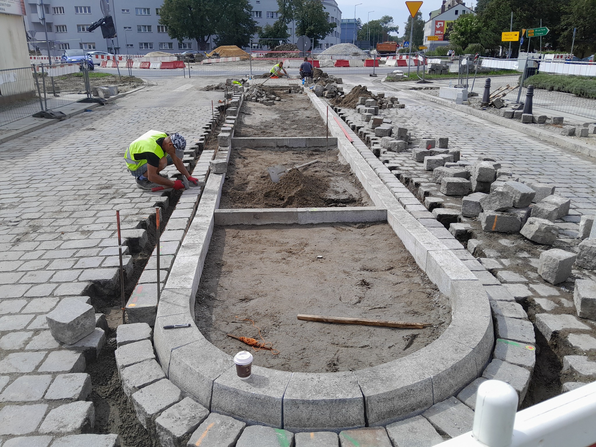 Układanie kostki brukowej na placu miejskim, widoczne przygotowane koryta, pracownicy w odblaskowych kamizelkach, porozrzucane kostki i kubek po kawie na obrzeżu.