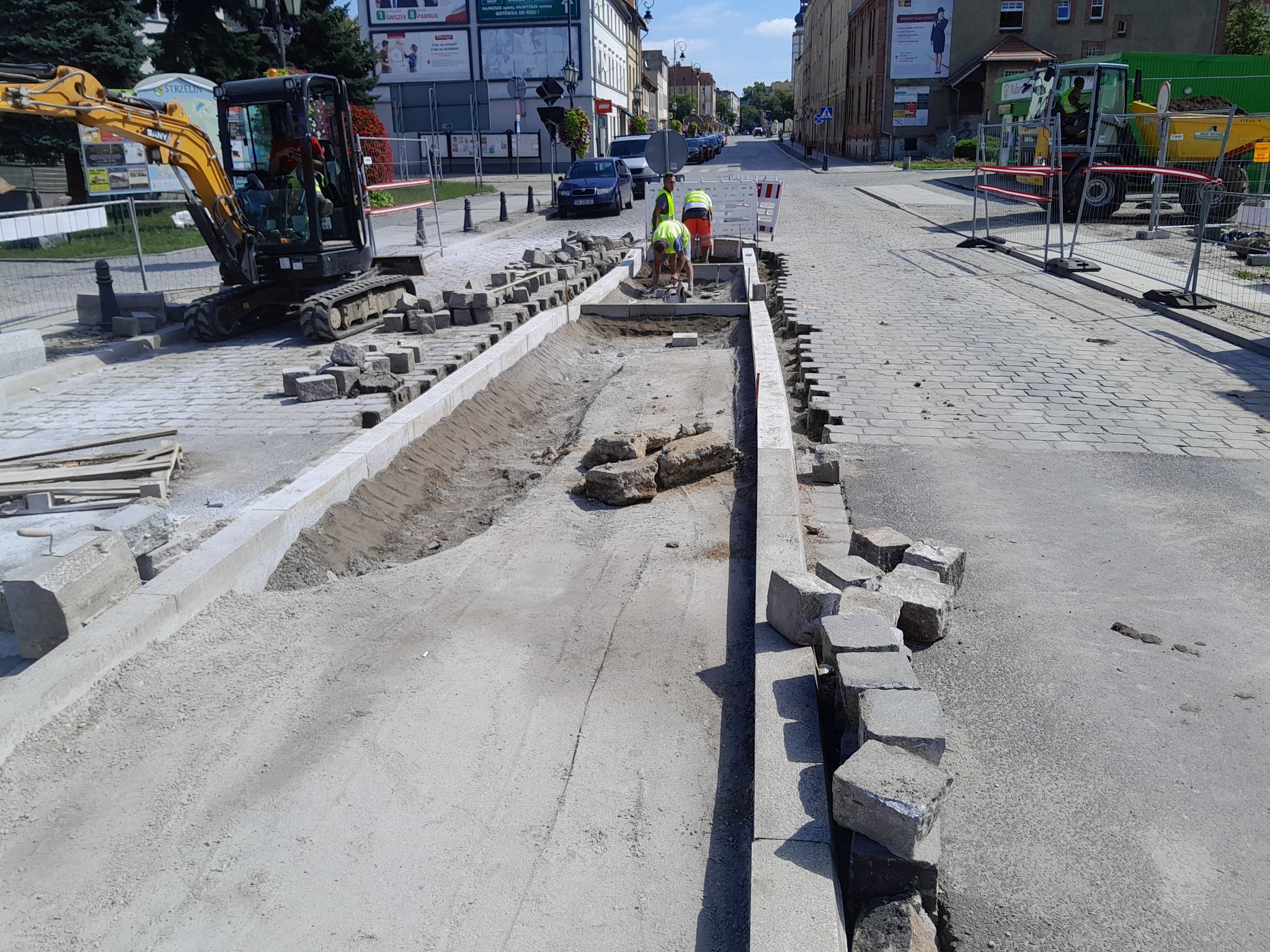 Prace brukarskie na ulicy w trakcie realizacji: wykop z ułożonymi krawężnikami, robotnicy w odblaskowych kamizelkach układają kostkę, w tle minikoparka i wywrotka.