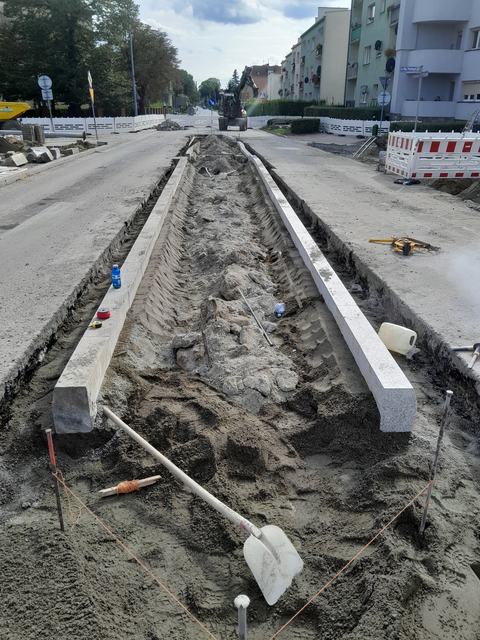 Przygotowanie podłoża pod drogę: wykop z ułożonymi obrzeżami chodnikowymi, widoczne narzędzia, piasek i w oddali maszyny budowlane na tle zabudowy miejskiej.