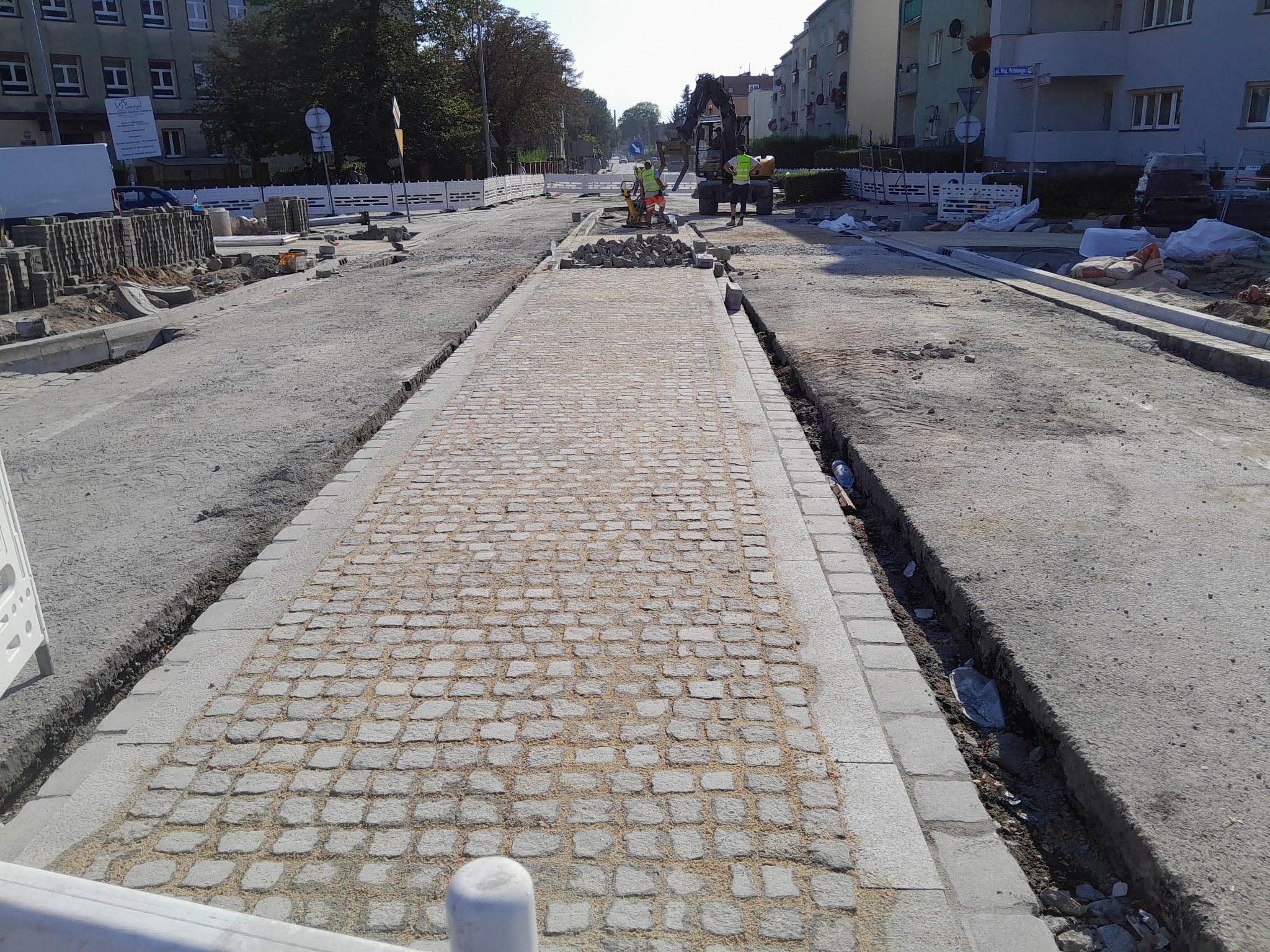 Układanie kostki brukowej na nowo budowanej drodze we Wrocławiu, z widocznym wykopem po bokach i pracownikami używającymi zagęszczarki w tle.