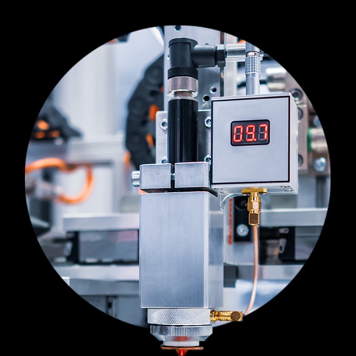 Precyzyjny detal głowicy lasera przemysłowego z wyświetlaczem, w srebrnej obudowie, okrągłe ujęcie na czarnym tle, widoczne przewody i elementy mocujące.