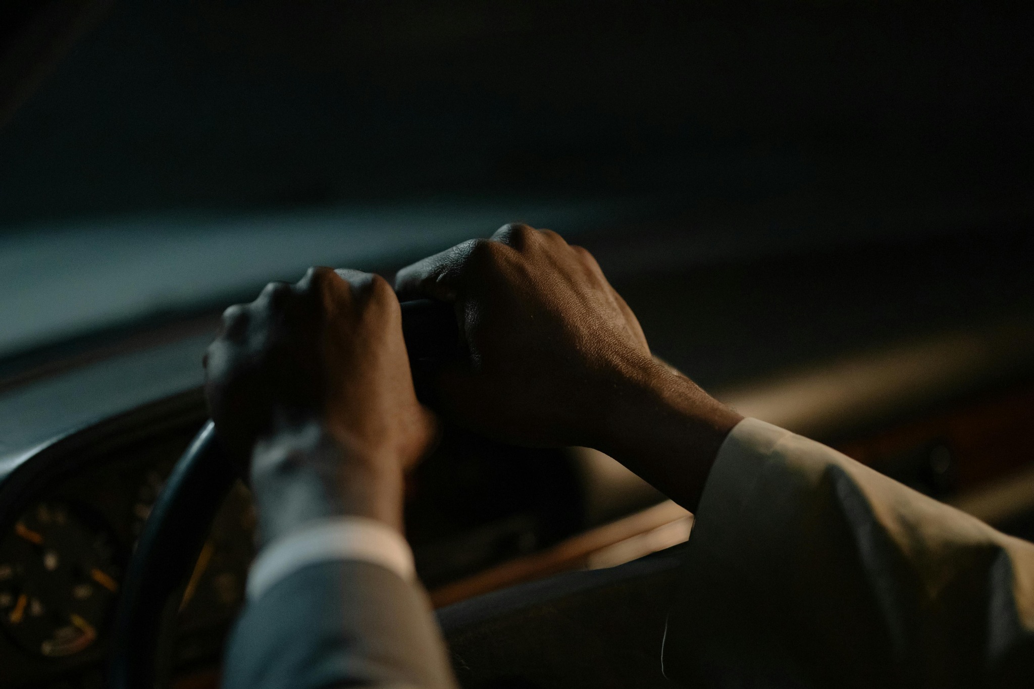 Dłonie kierowcy o ciemnej karnacji mocno trzymające kierownicę w ciemnym wnętrzu samochodu. Widoczny fragment deski rozdzielczej i rękaw marynarki.