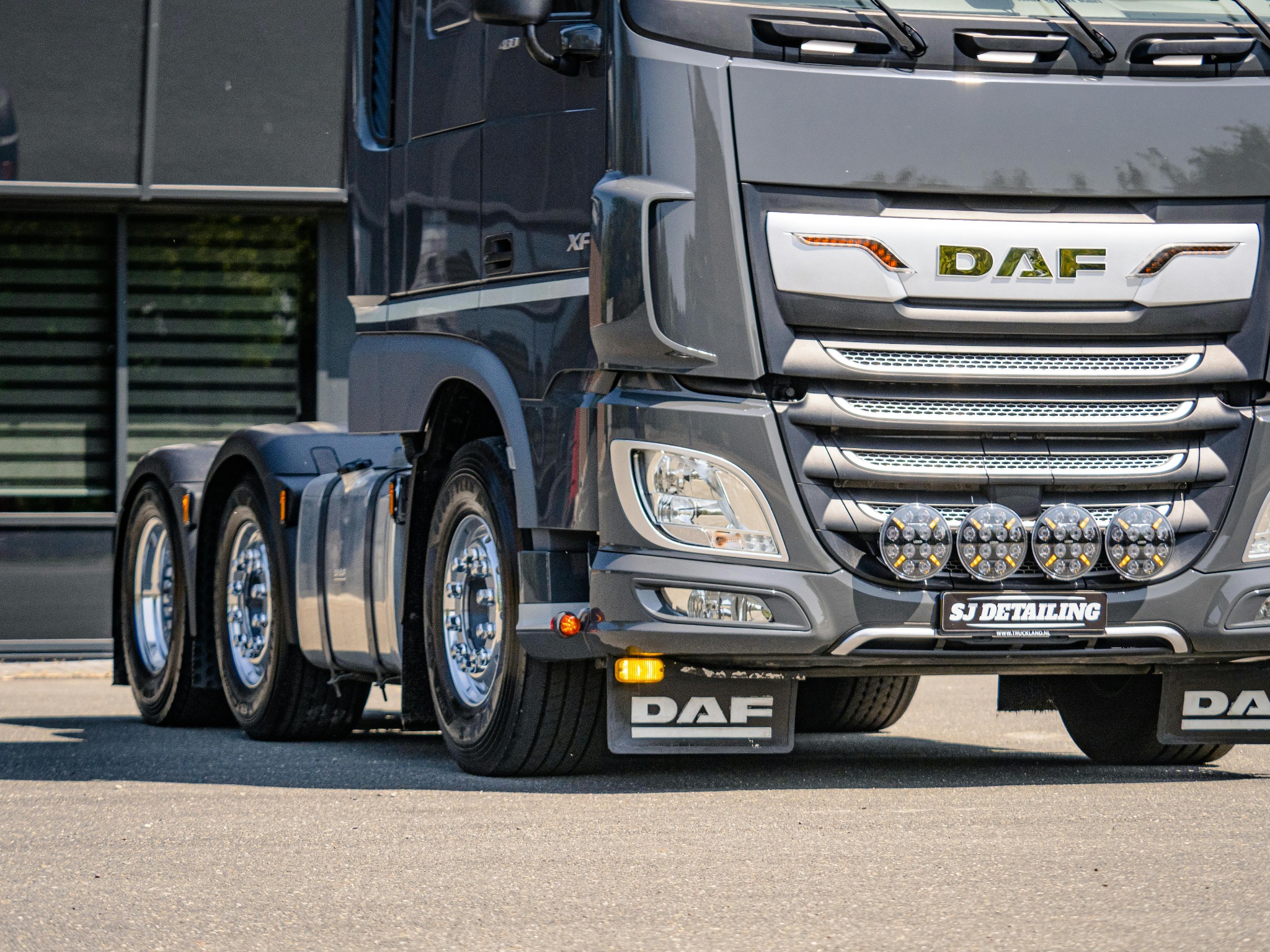 Szary ciągnik siodłowy DAF XF z dodatkowym oświetleniem LED i tabliczką 'SJ Detailing' na tle nowoczesnego budynku. Widok z przodu, niski kąt.