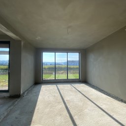 ProTynk - Wnętrze pokoju w stanie deweloperskim po nałożeniu gładzi, widoczne okno z widokiem na zewnątrz, surowy beton na podłodze i ściany przygotowane do dalszych prac wykończeniowych.