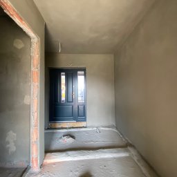 ProTynk - Korytarz w trakcie prac wykończeniowych: ściany i sufit pokryte świeżą warstwą szarej gładzi, widoczne surowe obramowanie drzwi po lewej stronie i ciemne drzwi wejściowe na wprost.