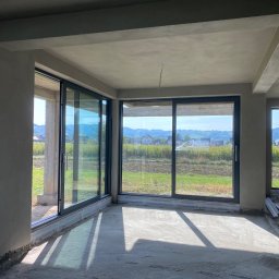 ProTynk - Surowe wnętrze w stanie deweloperskim z dużymi przesuwnymi oknami, szare ściany i sufit po wstępnym wykończeniu, widok na zielone pola i wzgórza za oknami.