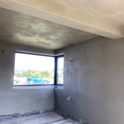 ProTynk - Pokój w trakcie prac wykończeniowych: świeżo nałożona szara warstwa na ścianach i suficie, widoczne wystające kable, okno z widokiem na zieleń i niebieski samochód.