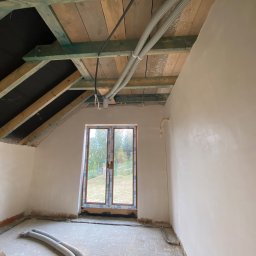 ProTynk - Poddasze w trakcie remontu: ściany po szpachlowaniu, widoczne rury instalacyjne pod drewnianym dachem z izolacją, okno z widokiem na zieleń.