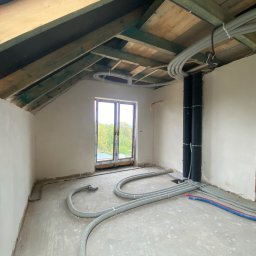 ProTynk - Poddasze w trakcie prac wykończeniowych: widoczne nieotynkowane ściany, drewniana konstrukcja dachu z poprowadzonymi przewodami wentylacyjnymi i elektrycznymi, okno z widokiem na zieleń.