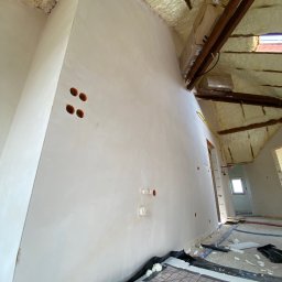 ProTynk - Ściana poddasza po nałożeniu gładzi, z widocznymi otworami na gniazdka elektryczne i instalacją ogrzewania podłogowego w trakcie montażu, drewniane belki stropowe z pianką izolacyjną i kartonami...