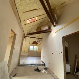 ProTynk - Poddasze w trakcie remontu: izolacja pianą PUR na skośnym dachu z widocznymi belkami, instalacja ogrzewania podłogowego, surowe ściany po szpachlowaniu z widocznymi punktami elektrycznymi...