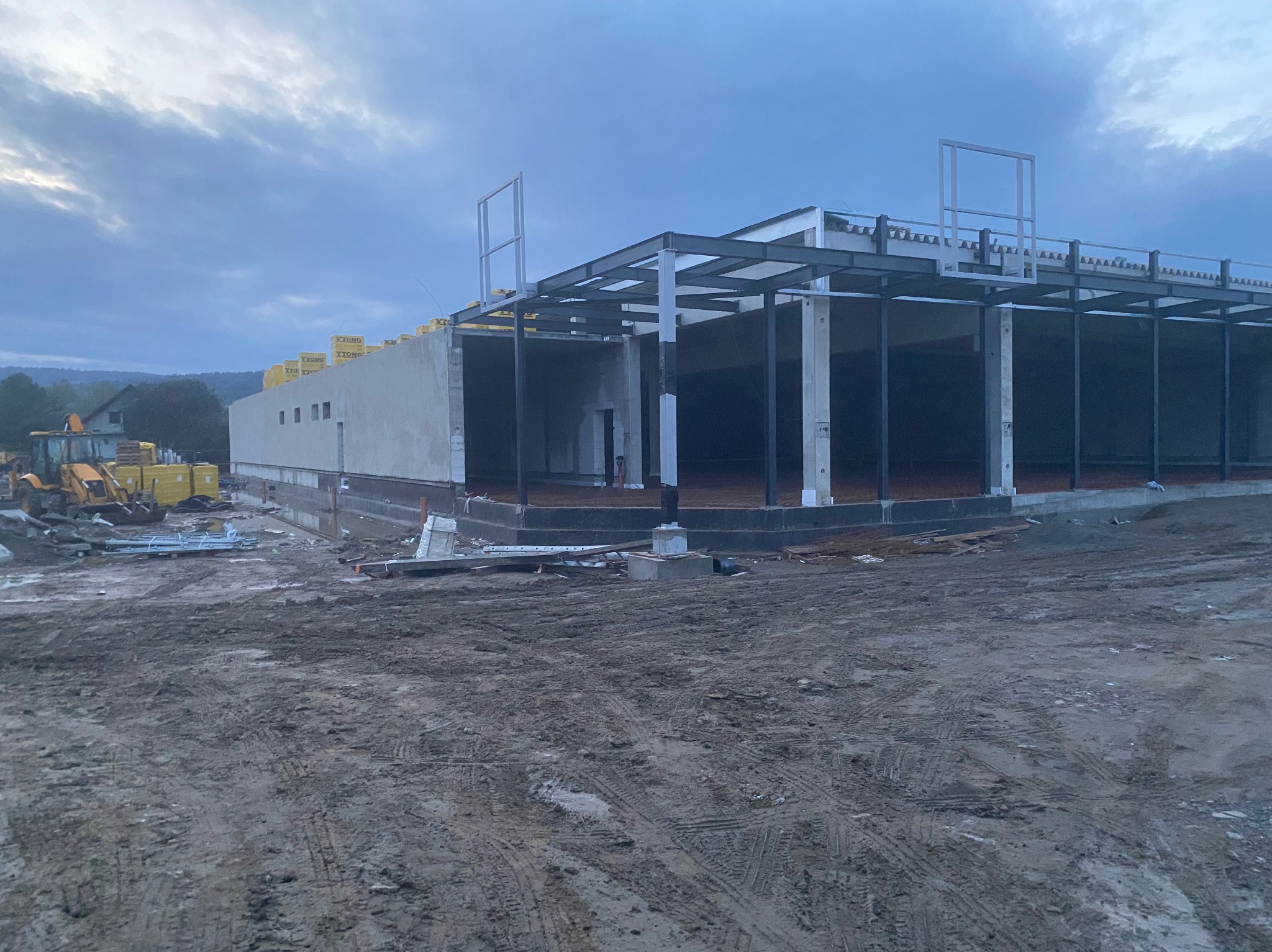 Budowa hali w stanie surowym otwartym z widoczną konstrukcją stalową, betonowymi słupami, częściowo wykończonymi ścianami z żółtymi płytami izolacyjnymi na dachu, oraz z ziemią wokół budynku.